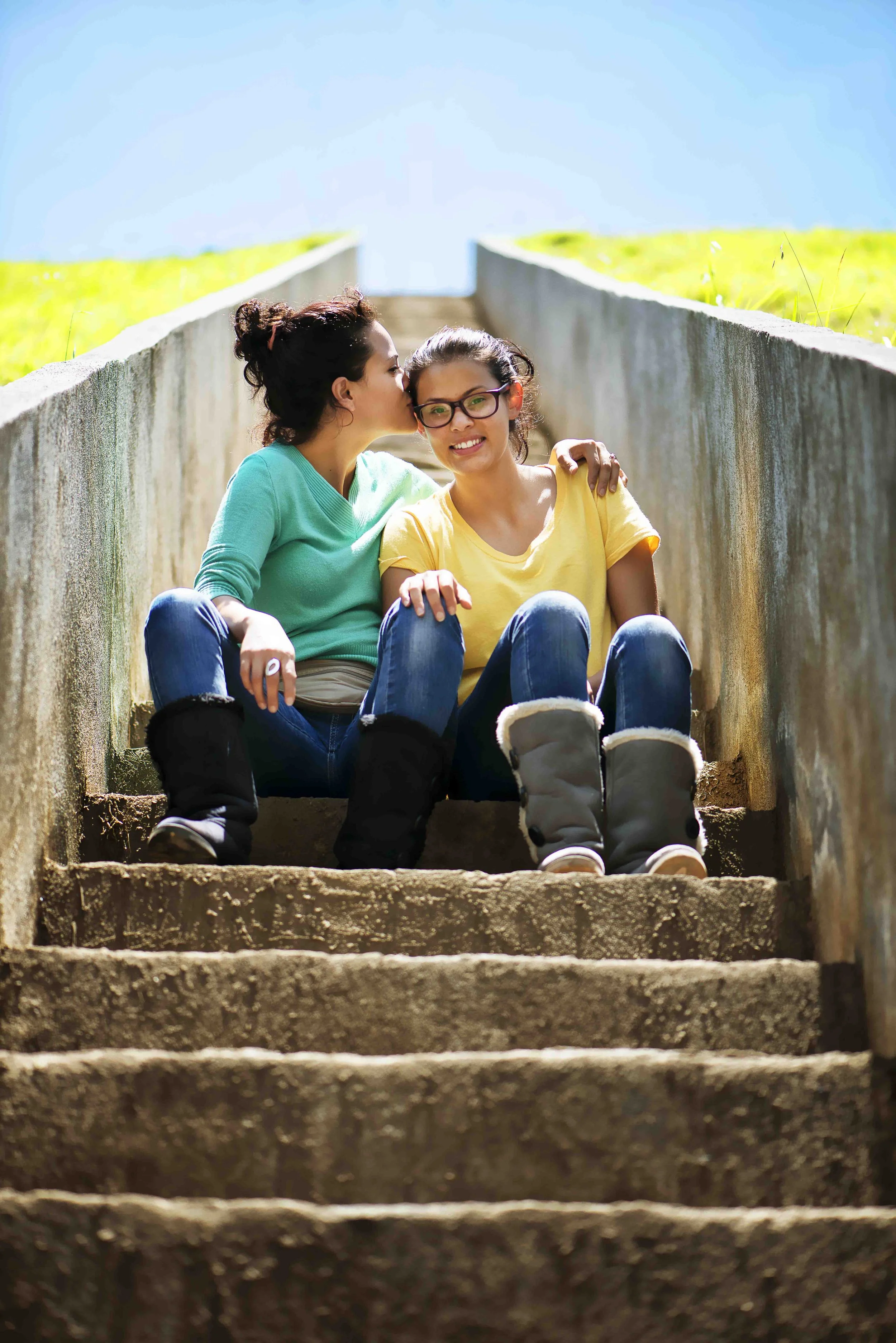 Two young women sitting on outdoor steps, one woman with glasses wearing a yellow shirt and the other woman kissing her on the cheek, wearing a teal shirt, both smiling.