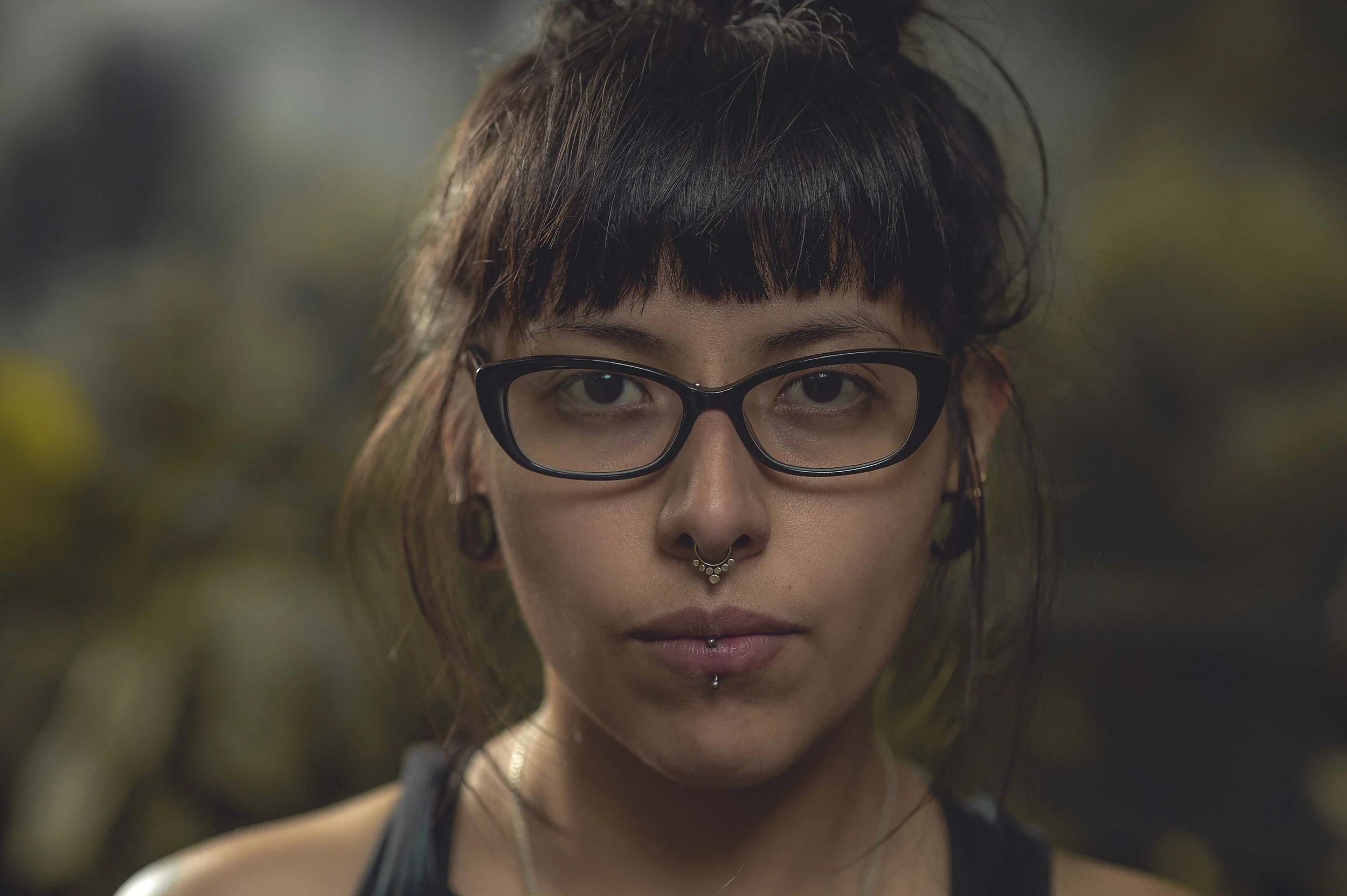 A woman with black hair, wearing glasses and multiple facial piercings, looks directly at the camera with a blurred outdoor background.