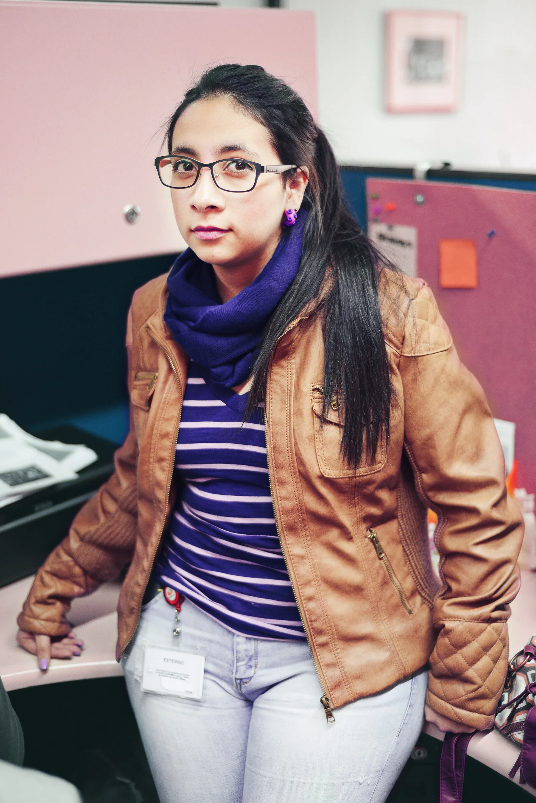 A woman with glasses, long dark hair, purple earrings, a purple scarf, a striped shirt, a tan jacket, and light-colored jeans, standing in an office.