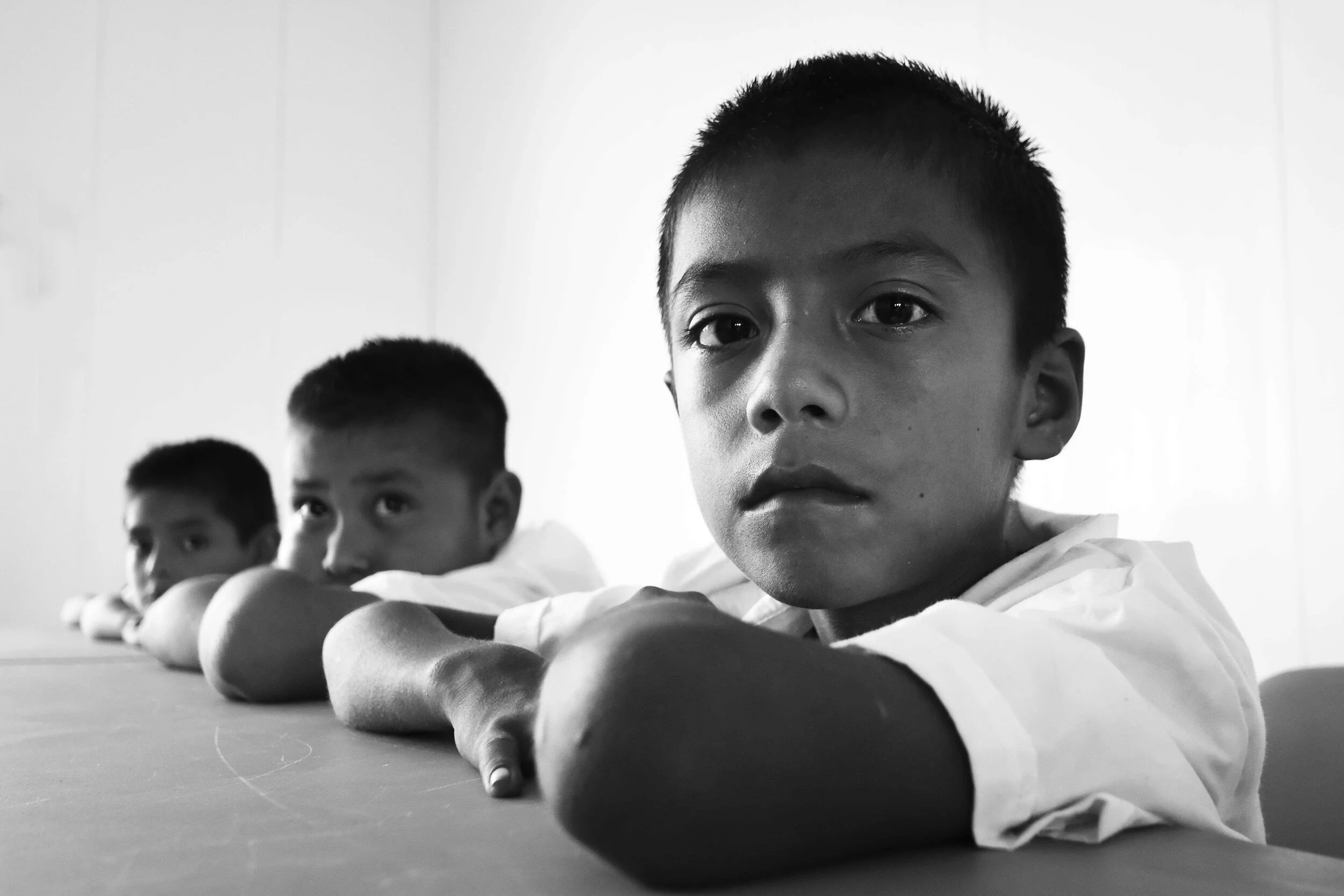Three boys lying on a desk with their arms extended forward, facing the camera in a classroom setting, black and white photograph.