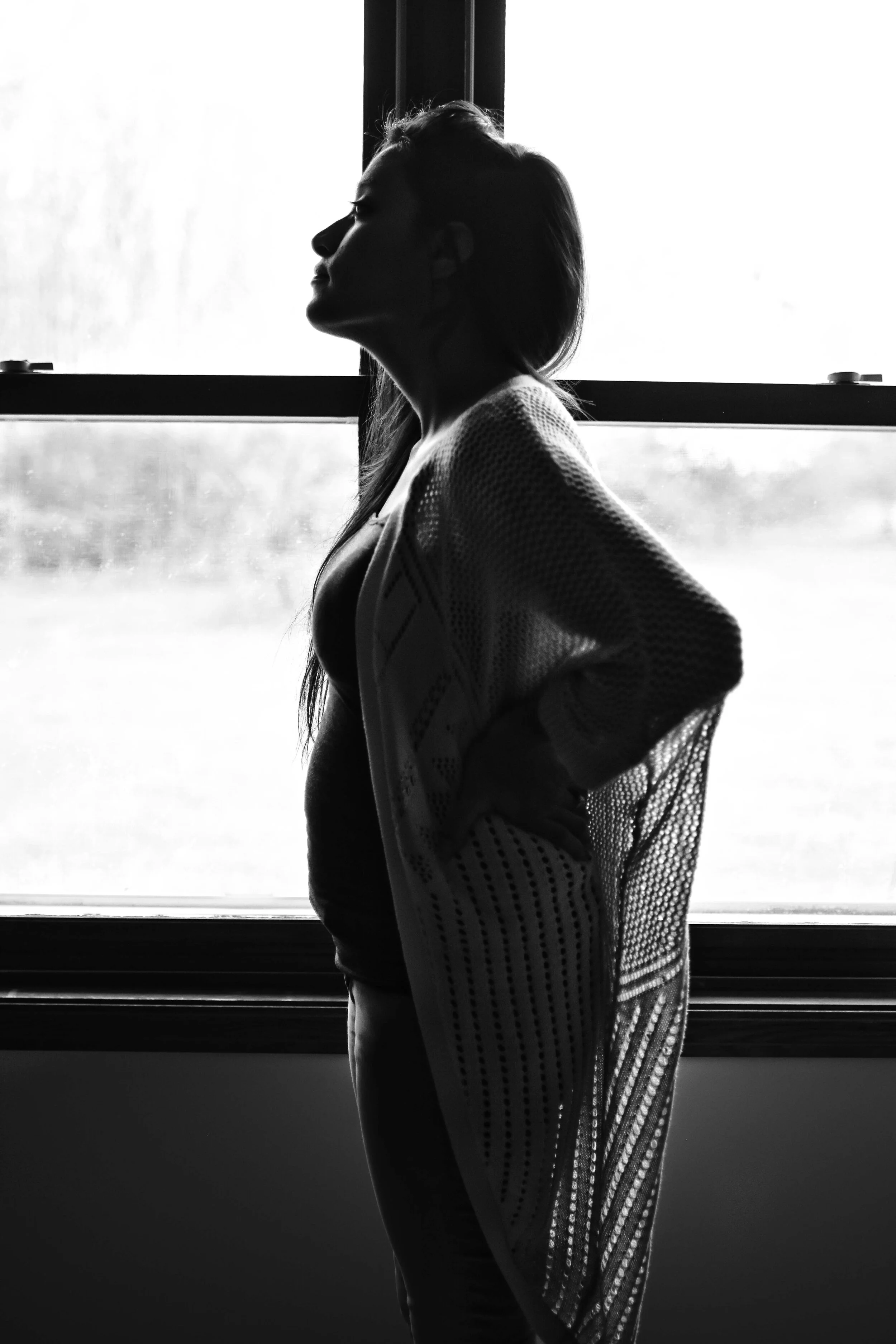 A woman in profile standing in front of a window, with natural light casting a silhouette. She has long hair, hands on her hips, and is wearing a loose, open-knit sweater.