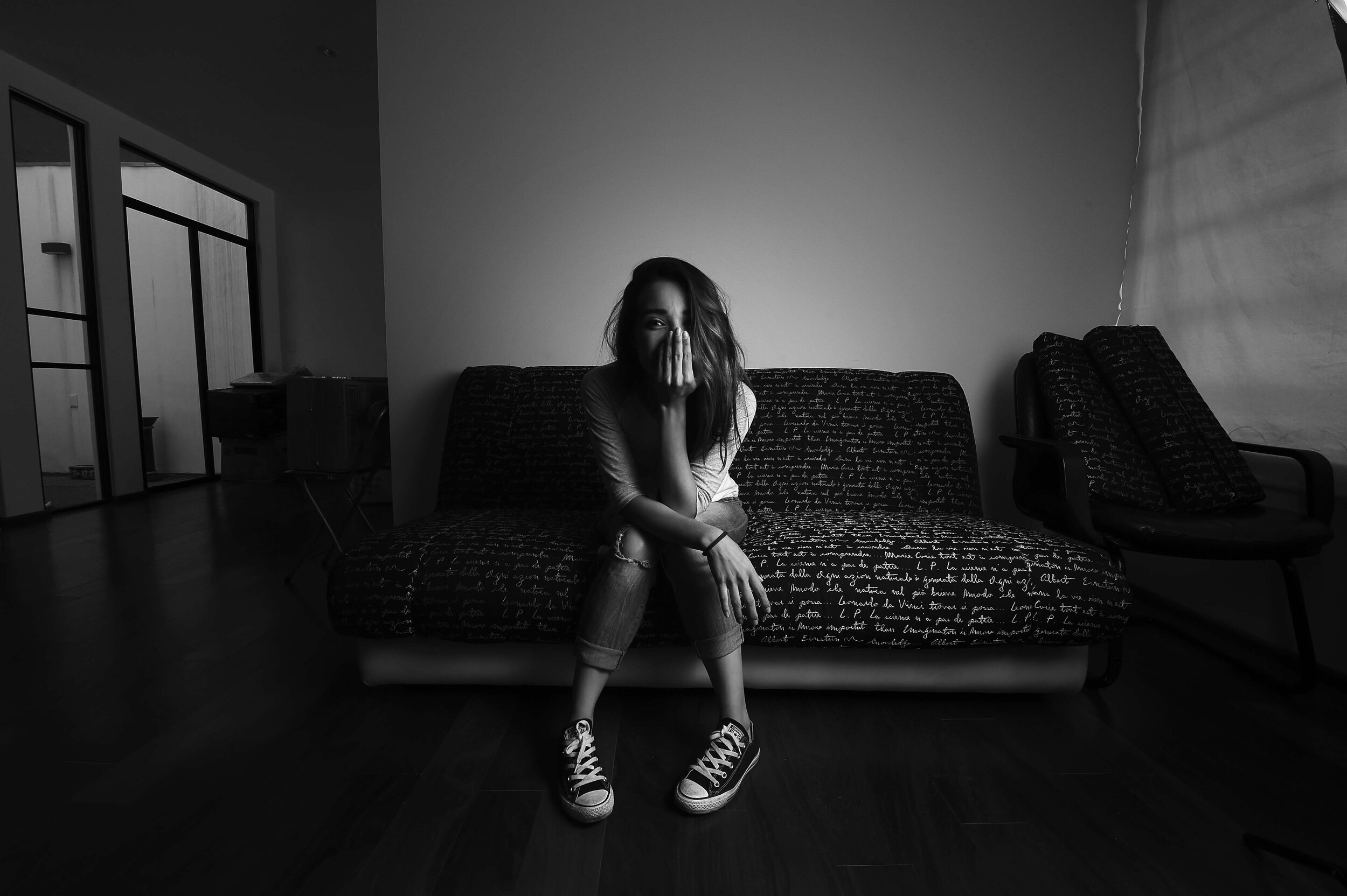 A young woman with long hair sitting on a patterned couch in a dimly lit room, covering her mouth with her hand, wearing sneakers and ripped jeans.