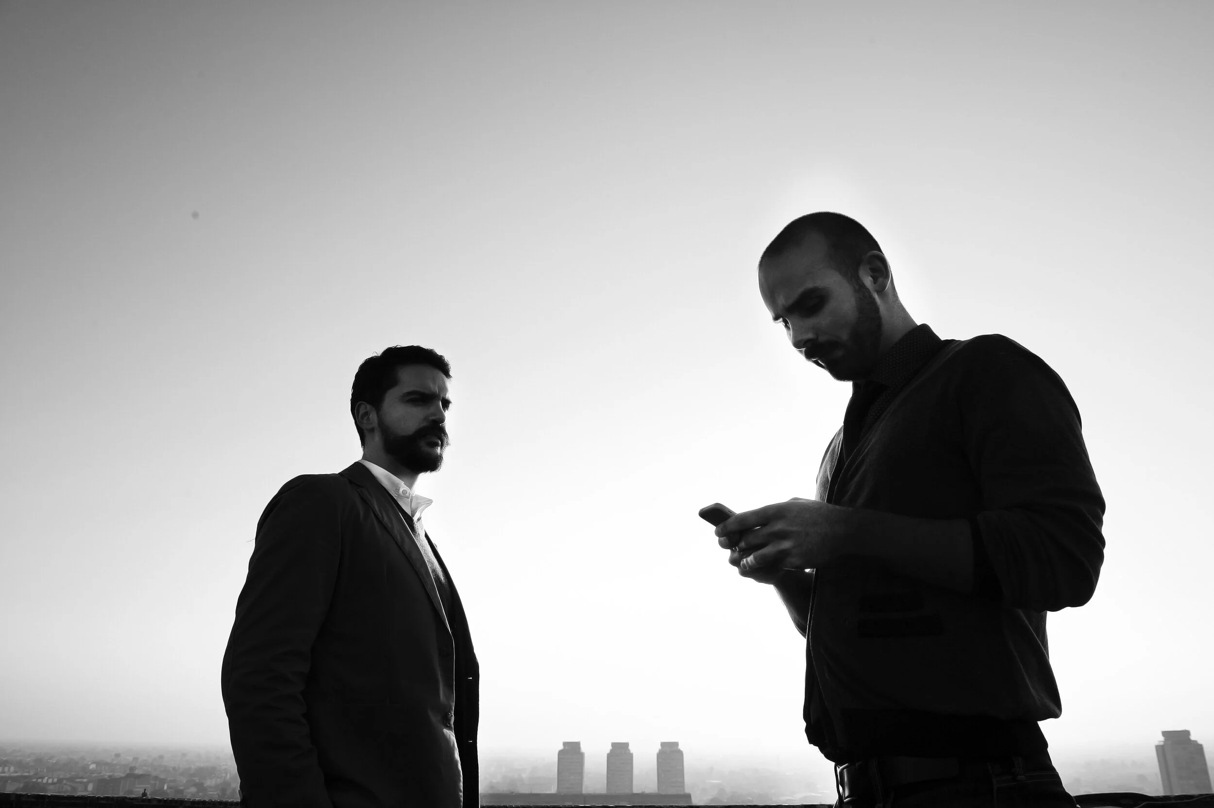 Silhouettes of two men standing outdoors with a cityscape in the background, one looking at his phone.