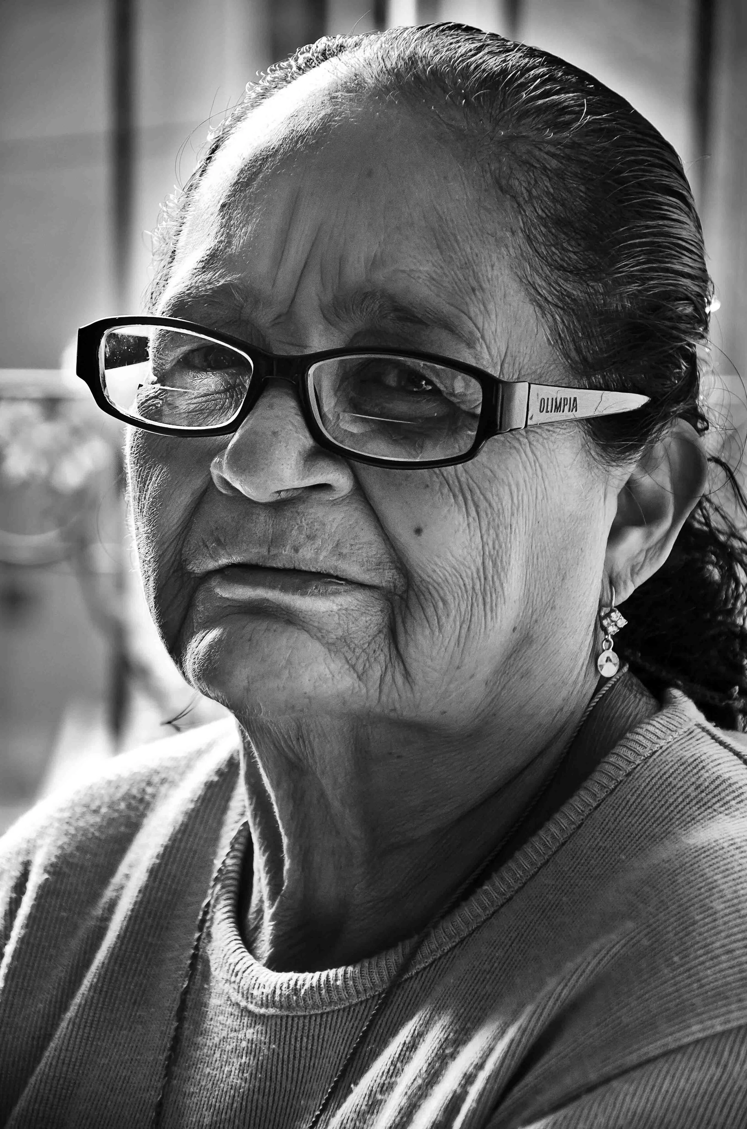 Close-up black-and-white portrait of an elderly woman with glasses, earrings, and dark hair slicked back.