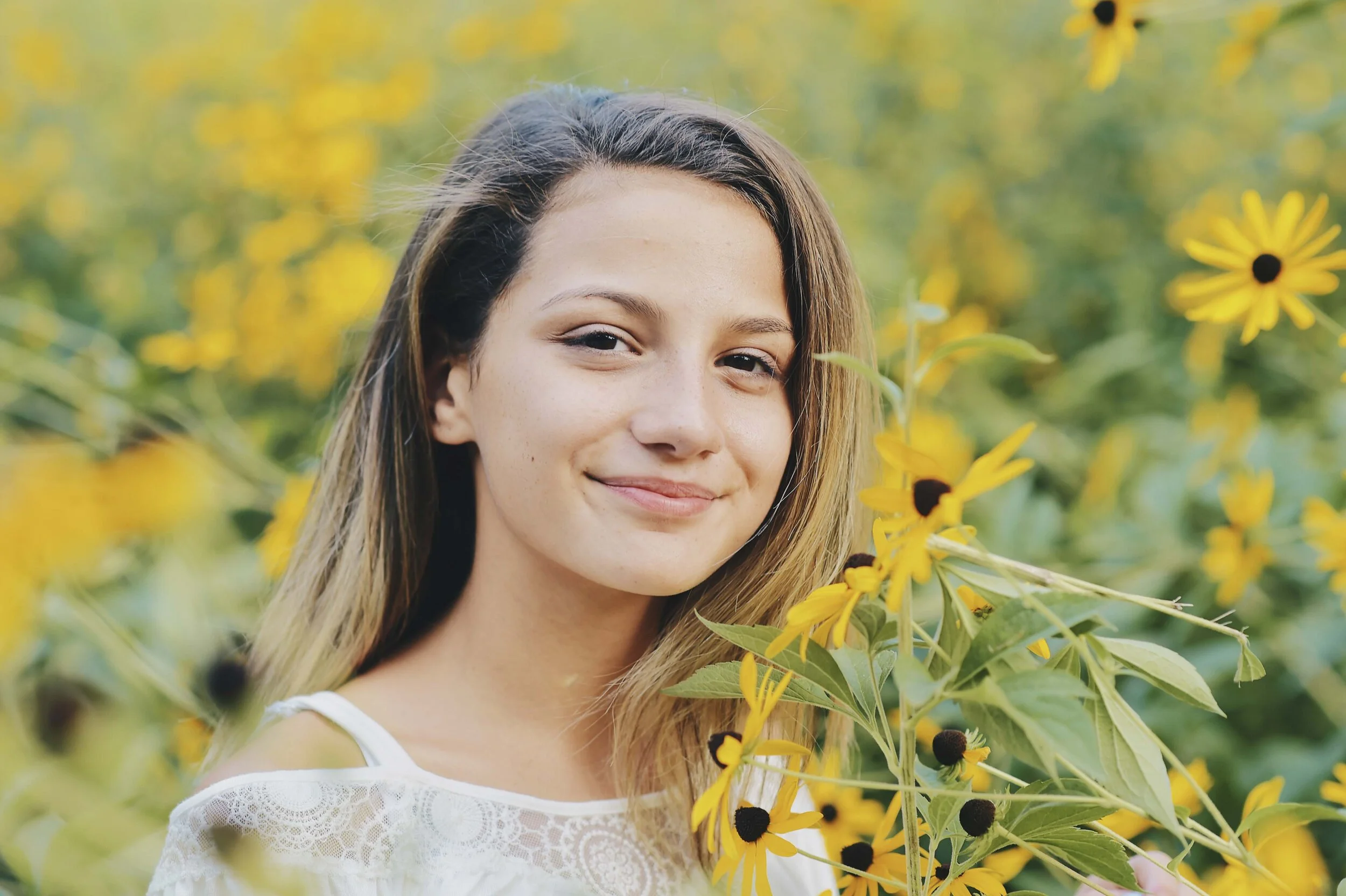 A young woman with long brown hair smiling in a field of yellow flowers with dark centers, wearing a white lace top.