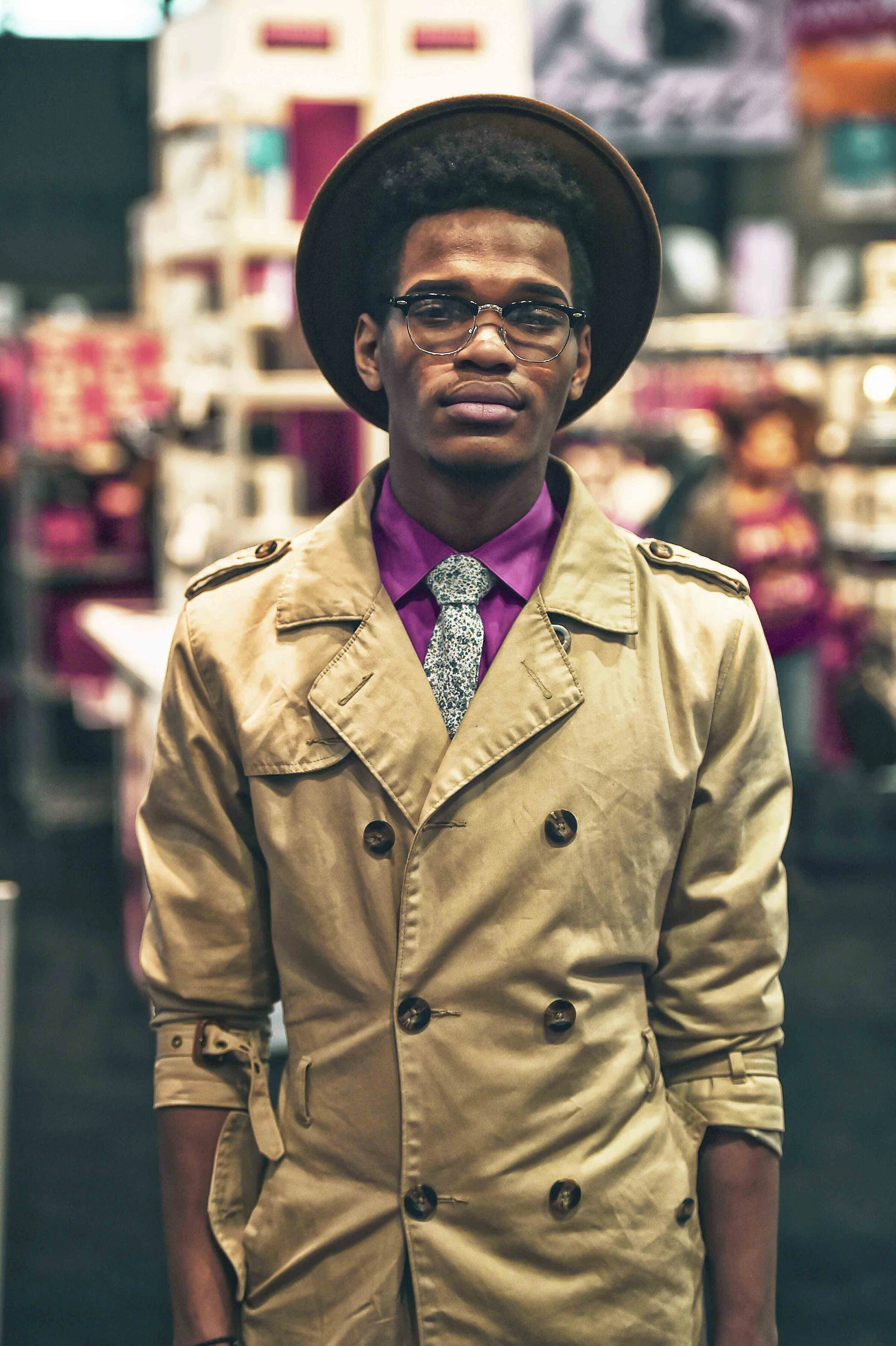 A young Black man wearing glasses, a black hat, a tan trench coat, a purple shirt, and a patterned tie stands in a store with blurred shelves and people in the background.