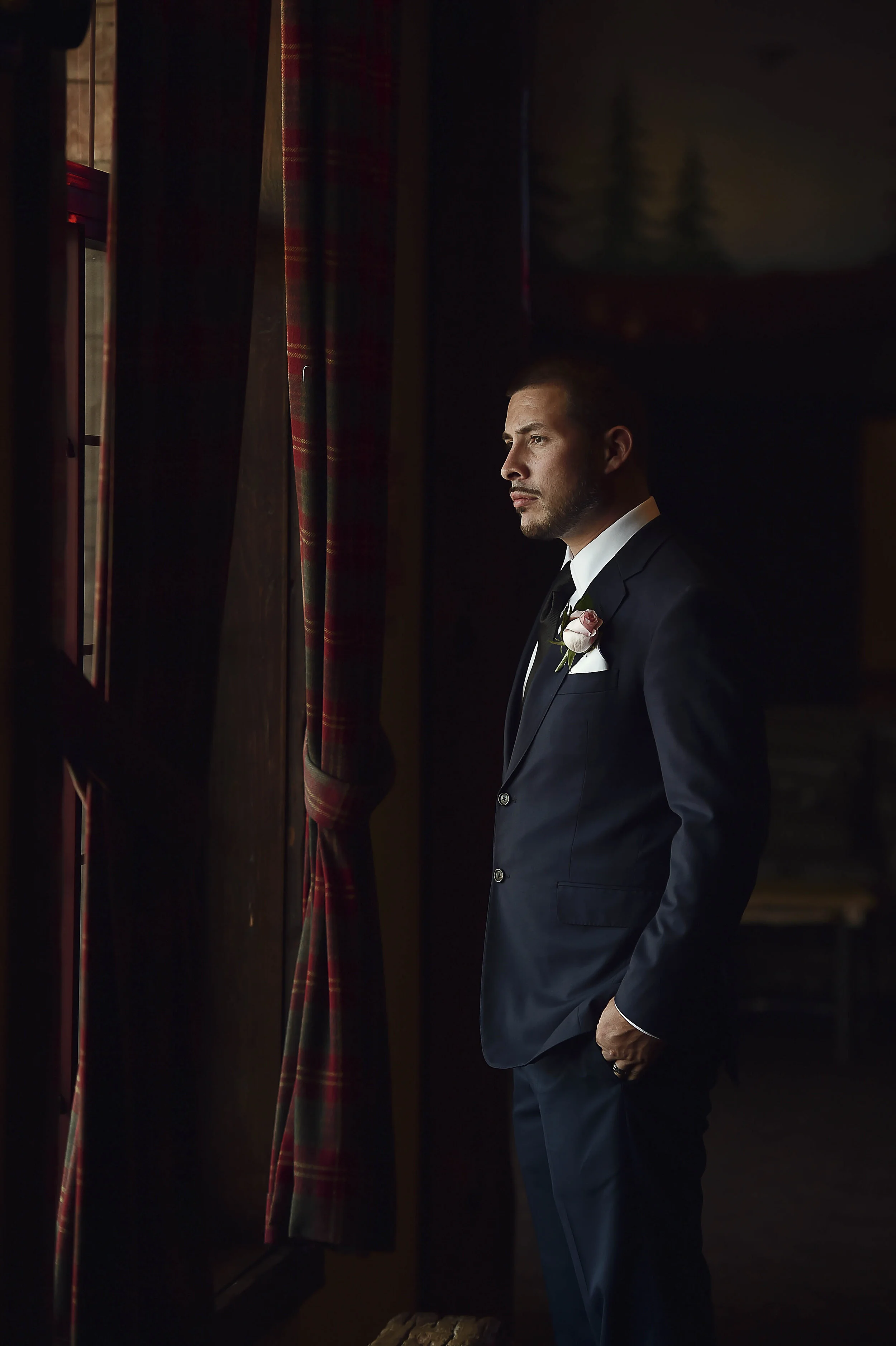 A man in a dark suit with a white shirt and a boutonniere stands by a window, looking outside in a dimly lit room.