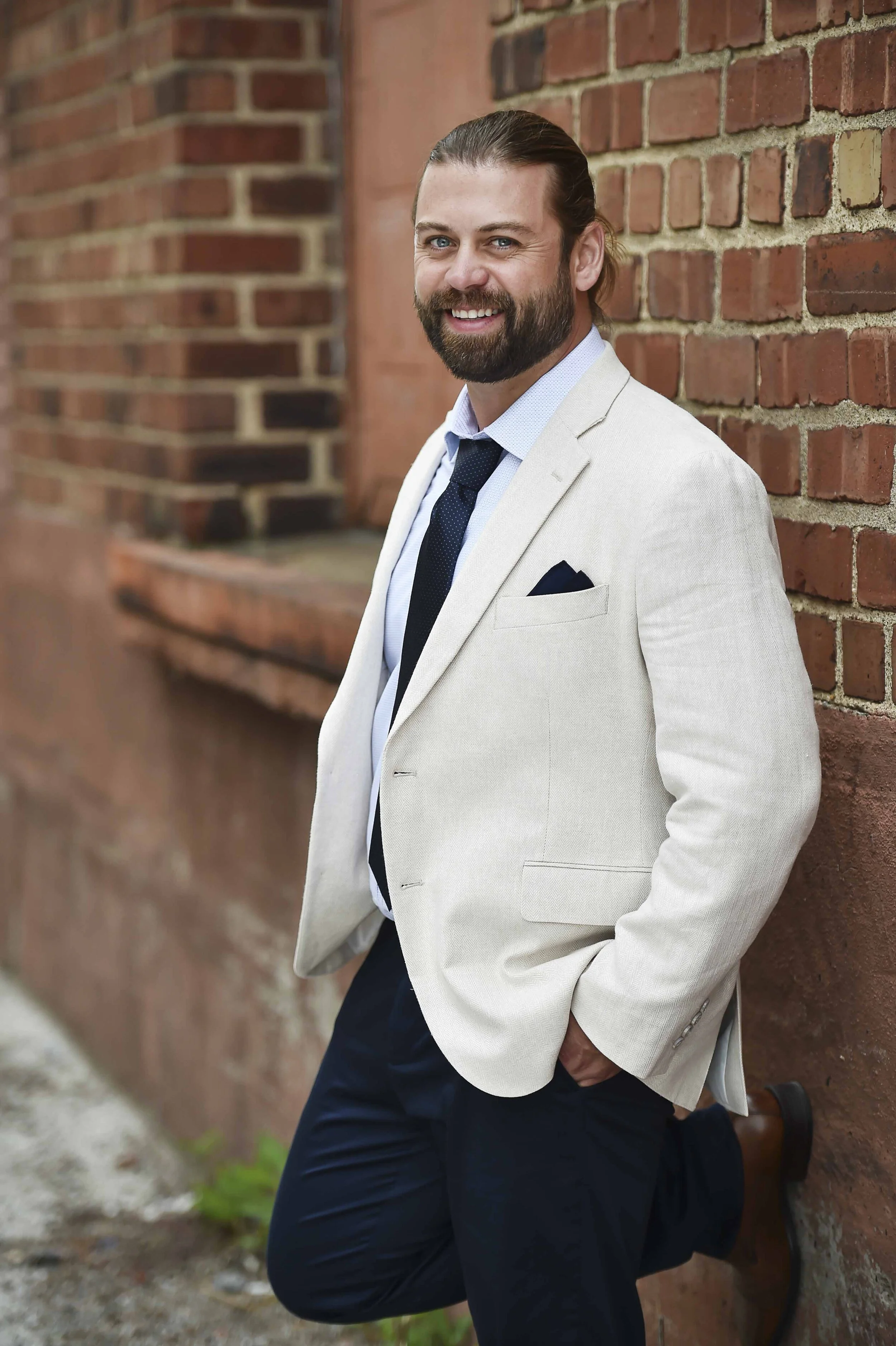 A man with a beard, wearing a light-colored blazer, blue dress shirt, dark tie, and dark pants, leaning against a brick wall with a smile.