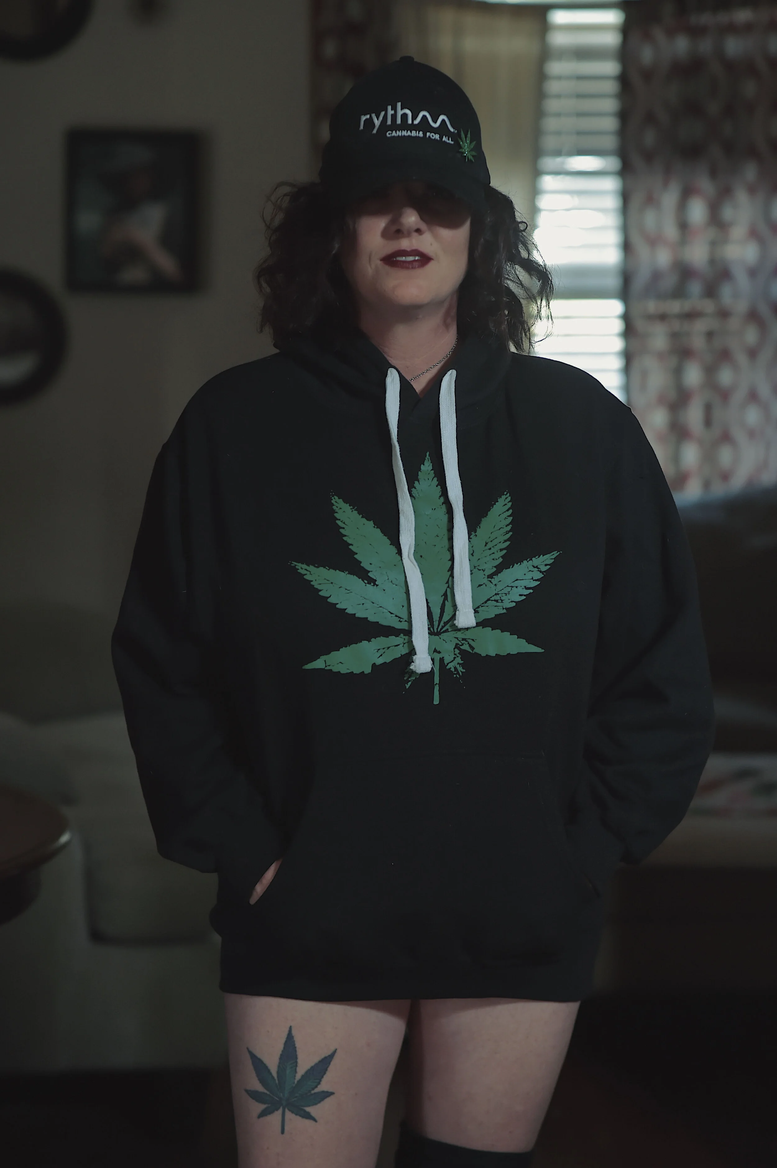 Woman standing indoors, wearing a black hoodie with a green cannabis leaf graphic on the front, a black cap with similar cannabis leaf and text, and showing a matching cannabis leaf tattoo on her upper thigh.