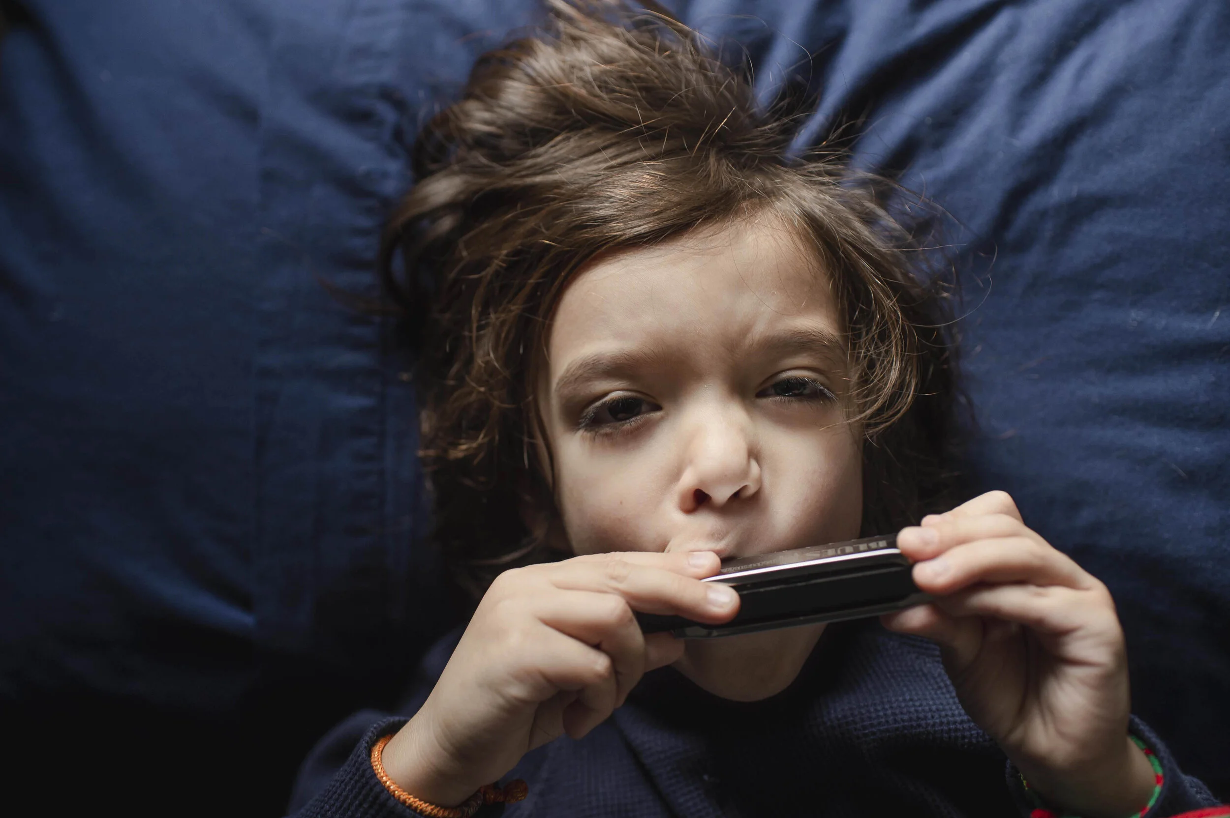 Young boy with brown hair lying on a dark blue pillow, holding a remote control close to his mouth, looking directly at the camera with a neutral expression.