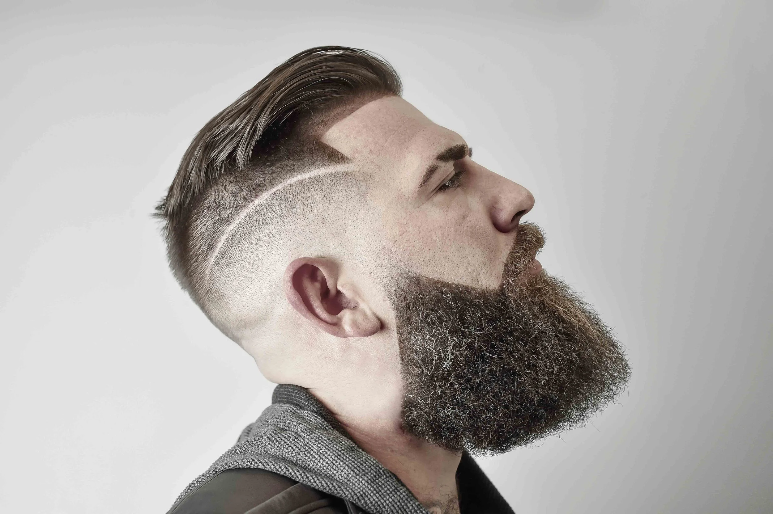 Side view of a man with a styled haircut and full beard, wearing a hoodie, against a plain background.