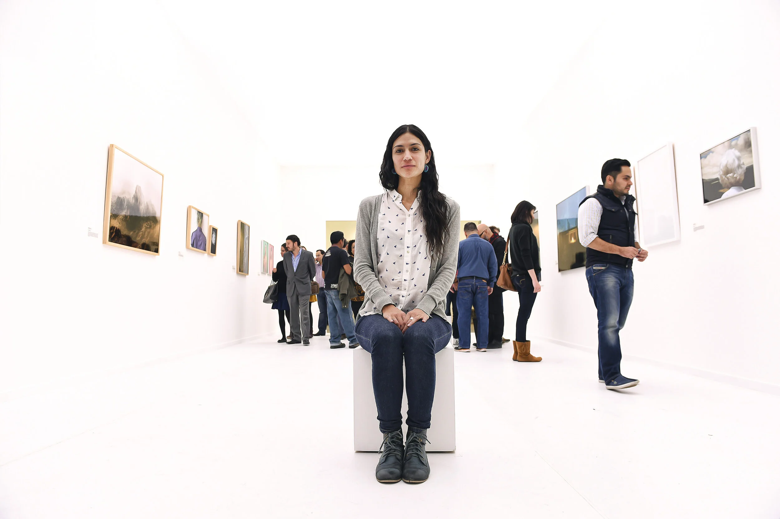 A woman sitting on a white box in an art gallery, surrounded by visitors viewing various paintings on white walls.