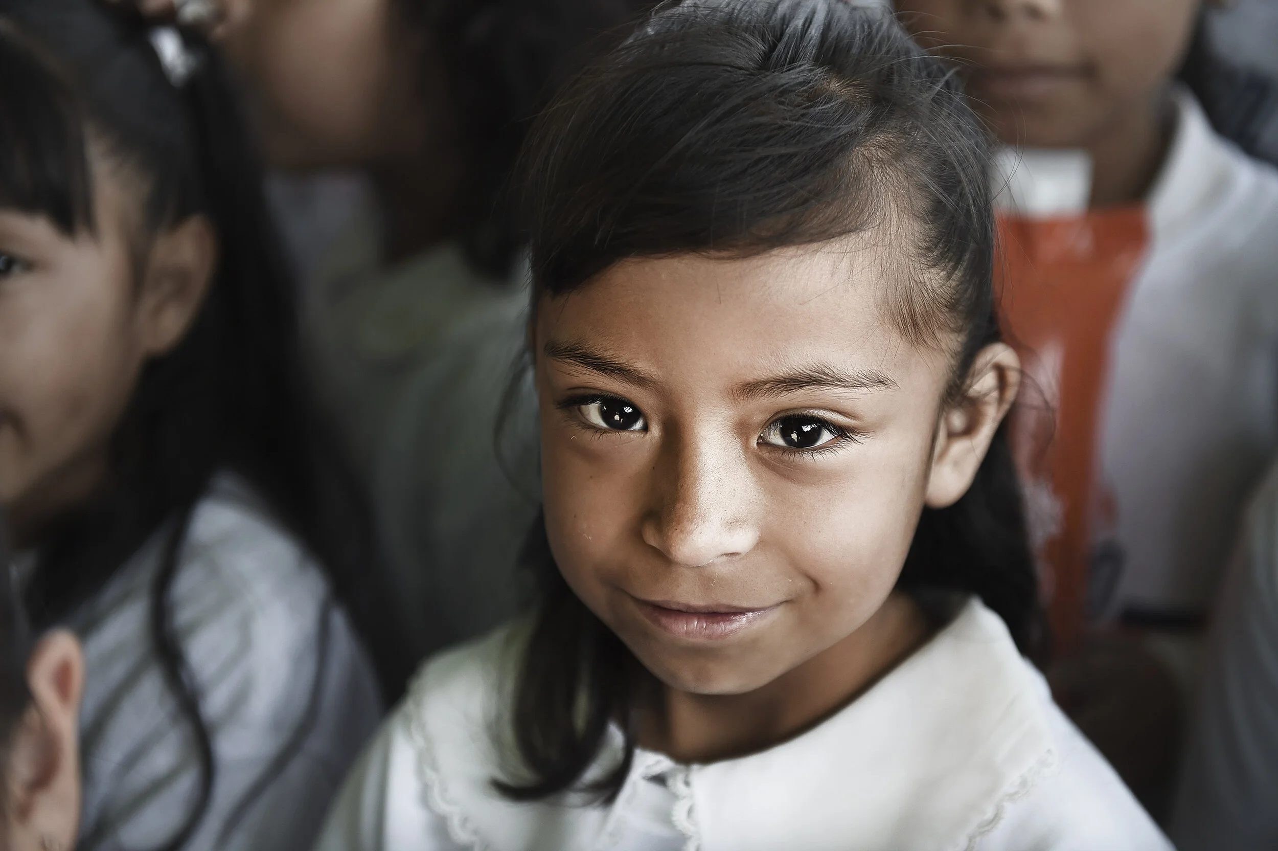 Close-up of a young girl with dark hair and brown eyes, smiling gently, surrounded by other children.