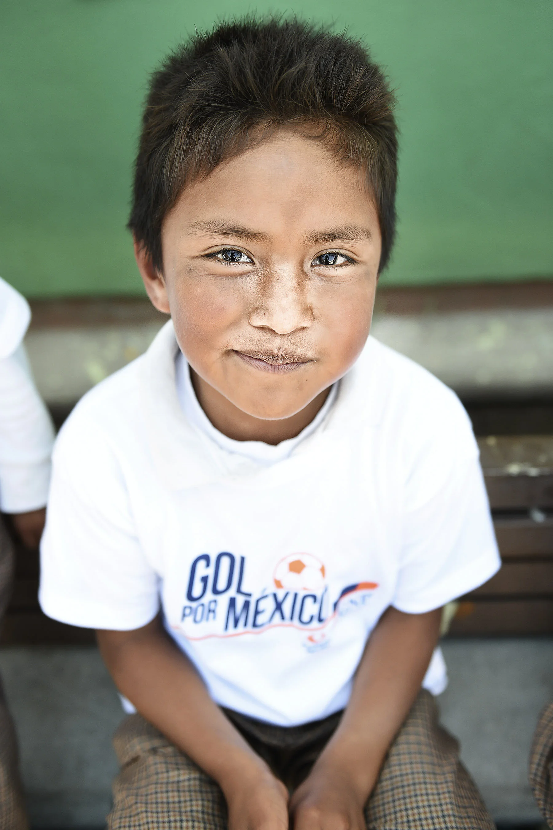 A young boy with dark hair and blue eyes, wearing a white T-shirt with 'GOL POR MEXICO' printed on it, sitting on a bench, looking up at the camera.