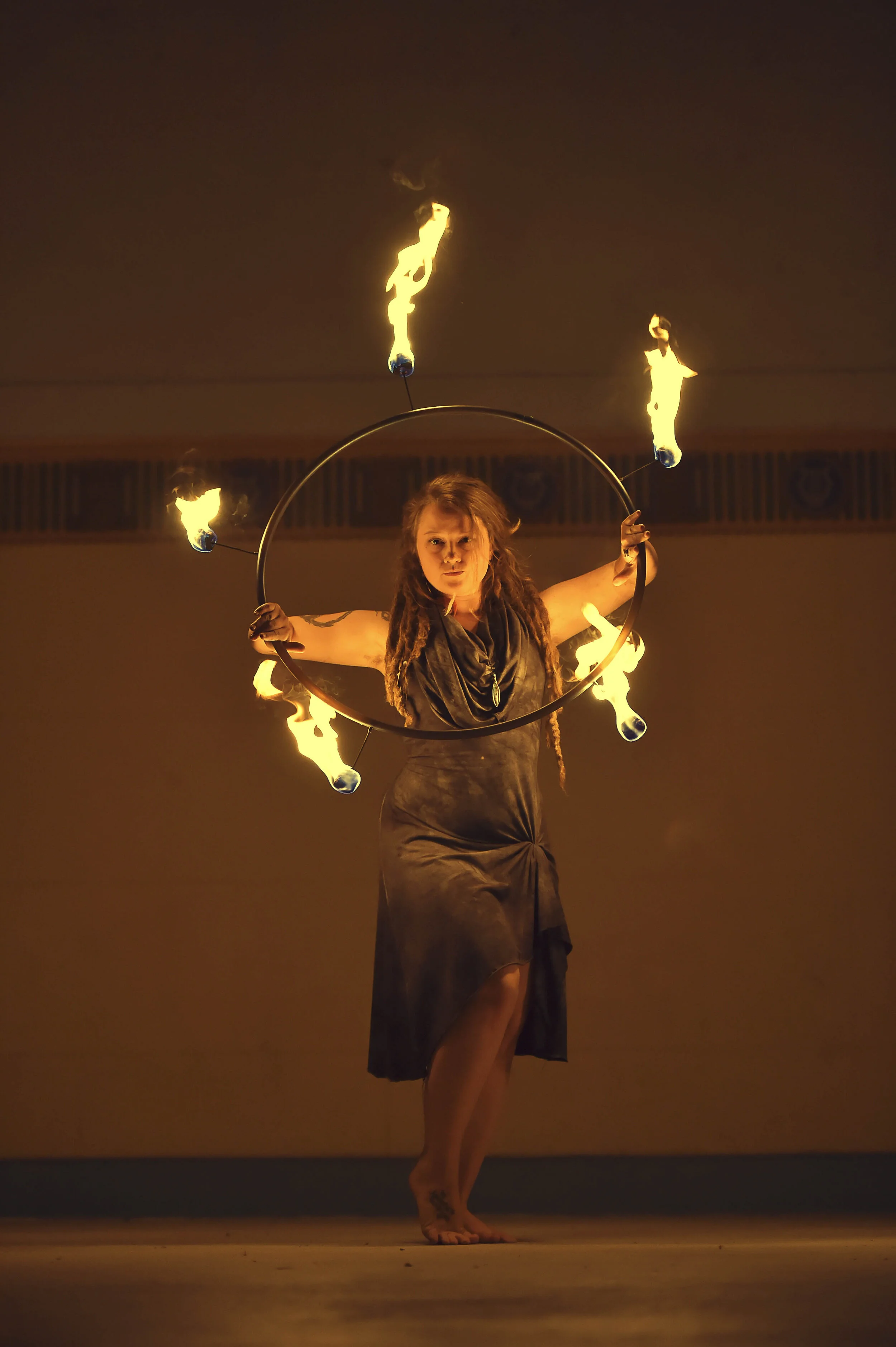 A female fire performer with dreadlocks is spinning a large metal hoop with flames on it, creating a fiery display during a performance.