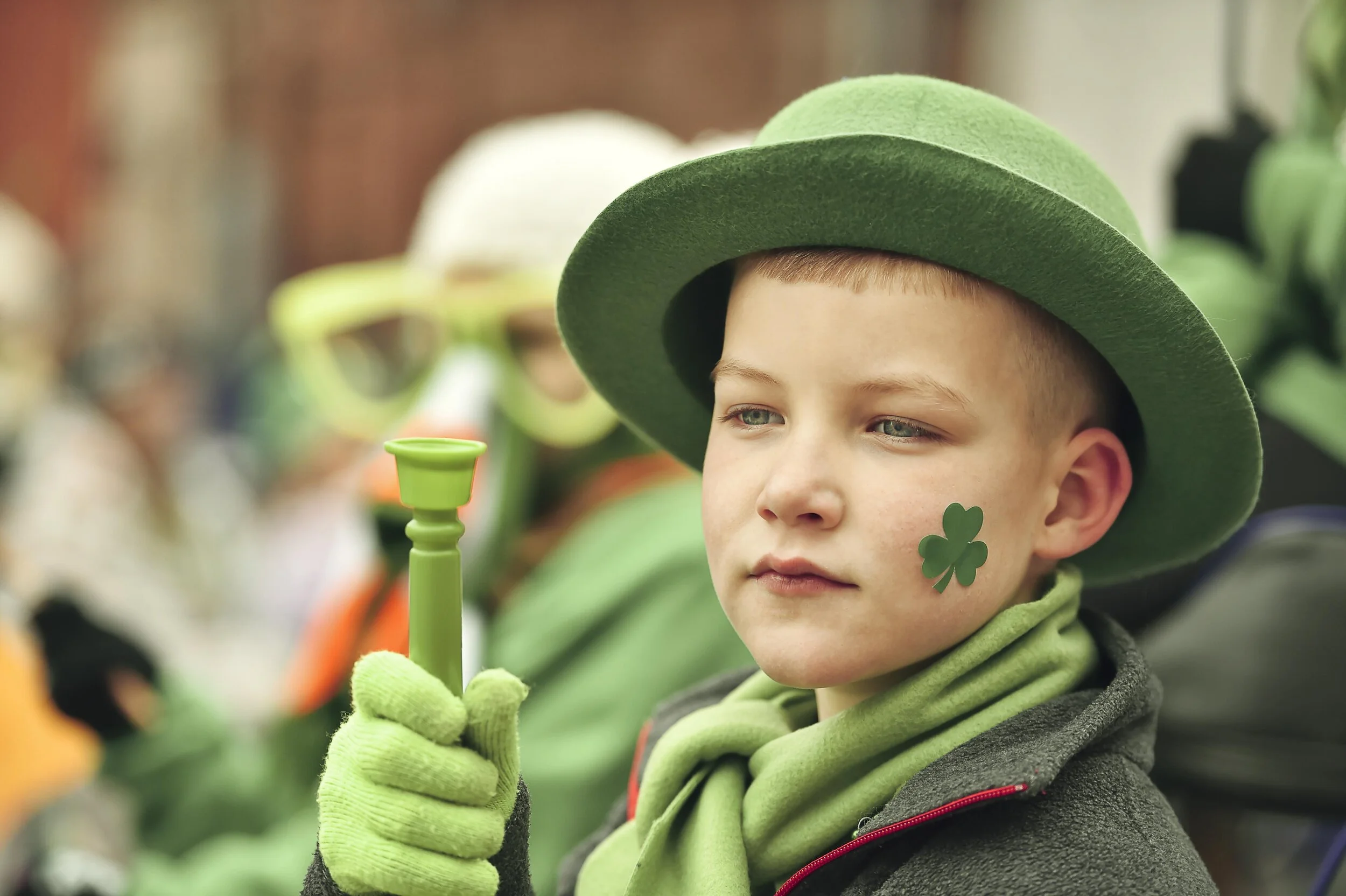 A young boy dressed in green, wearing a green hat, scarf, and gloves, holding a small green candle holder, with a shamrock sticker on his cheek, during a St. Patrick's Day celebration.