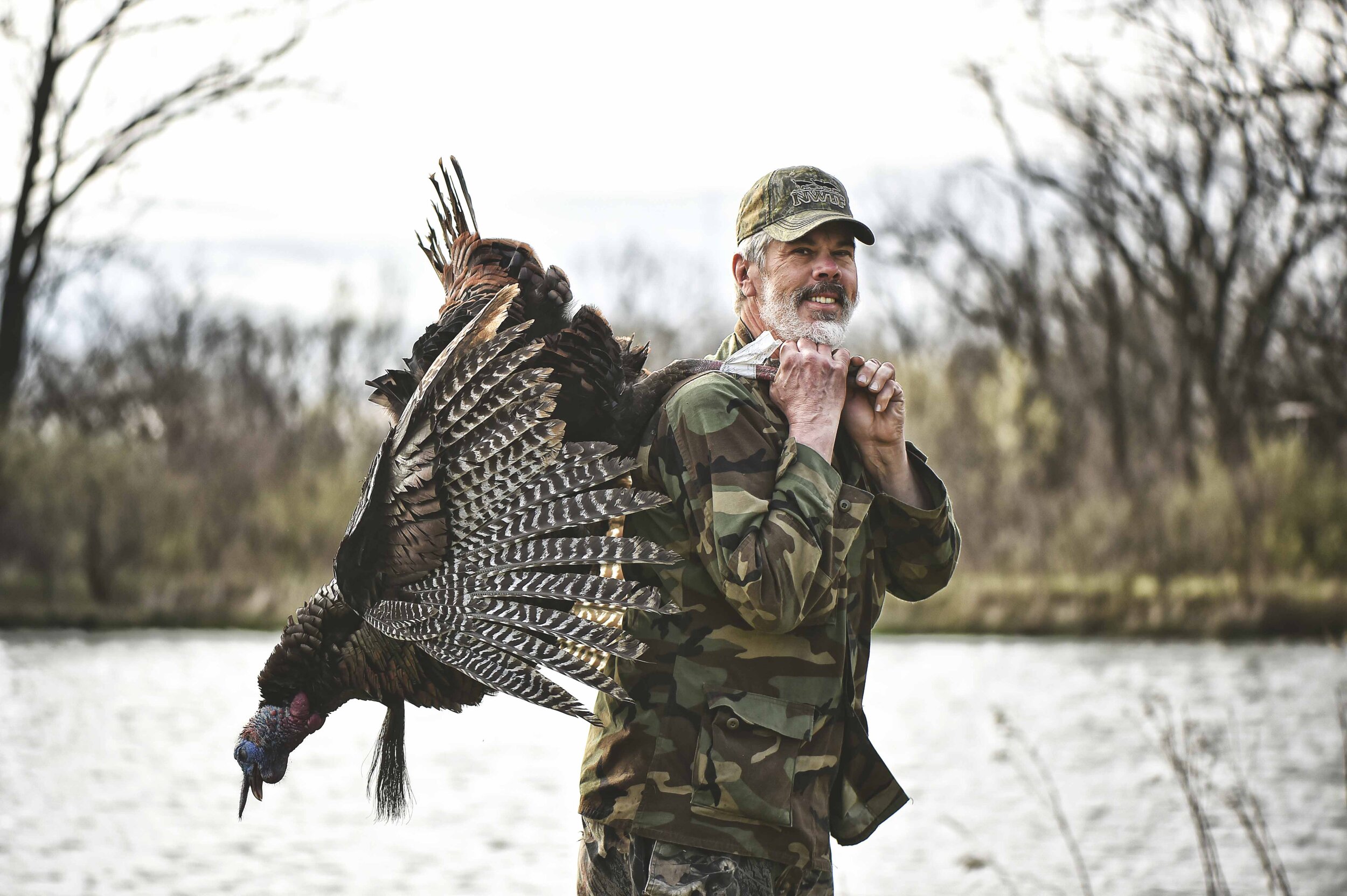 A man dressed in camouflage holding a large turkey over his shoulder outdoors near a body of water.