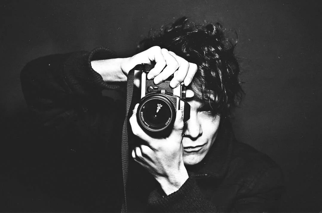Black and white photo of a person with curly hair holding a camera up to their face, partially covering one eye, against a dark background.