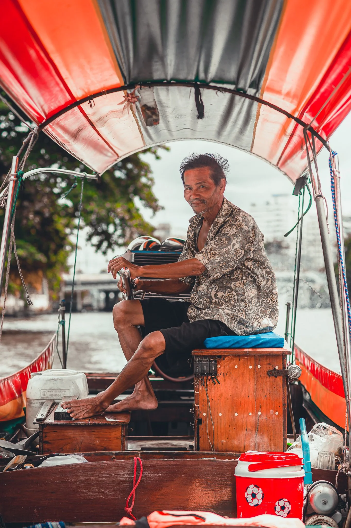 An older man sitting on a wooden seat on a boat, steering with his right hand, looking at the camera. The boat has a colorful canopy, and there are various items including a red cooler with soccer ball designs and other objects around him. Water and 