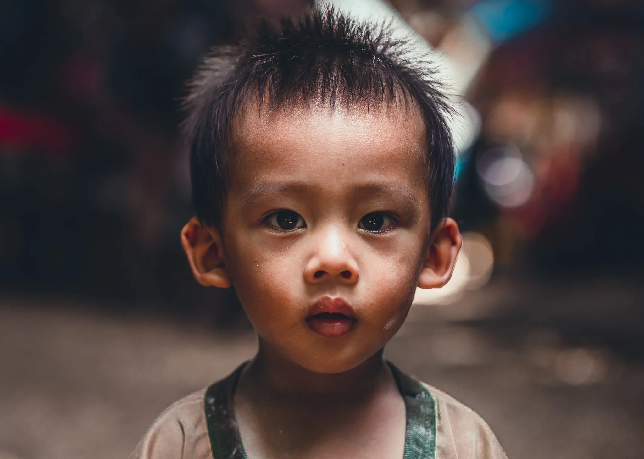 Close-up of a young boy with short black hair, dark eyes, and tan skin, standing outdoors with a blurred background.