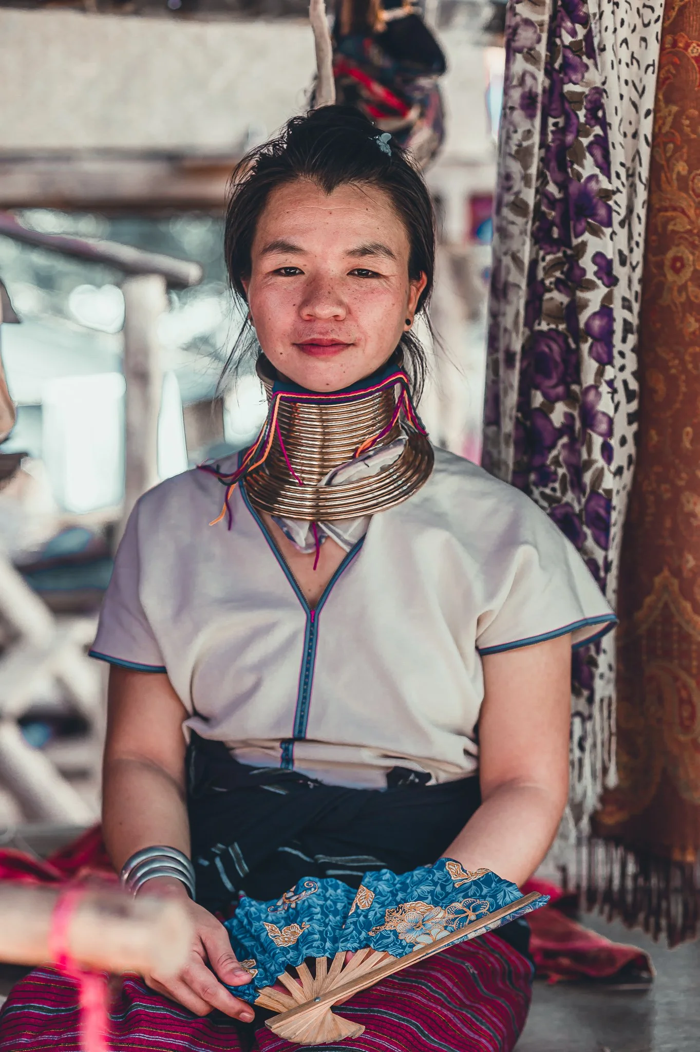 A woman wearing traditional attire with a large coil of brass rings around her neck, sitting indoors with patterned curtains in the background, holding a decorative fan.