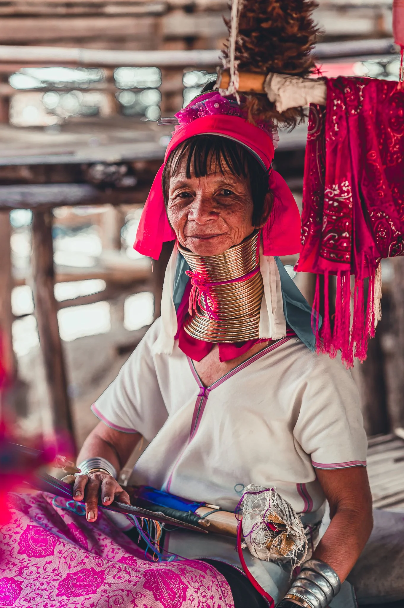 A woman from the Karen ethnic group wearing traditional brass neck rings and pink headgear, sitting and weaving textile in a rustic wooden setting.