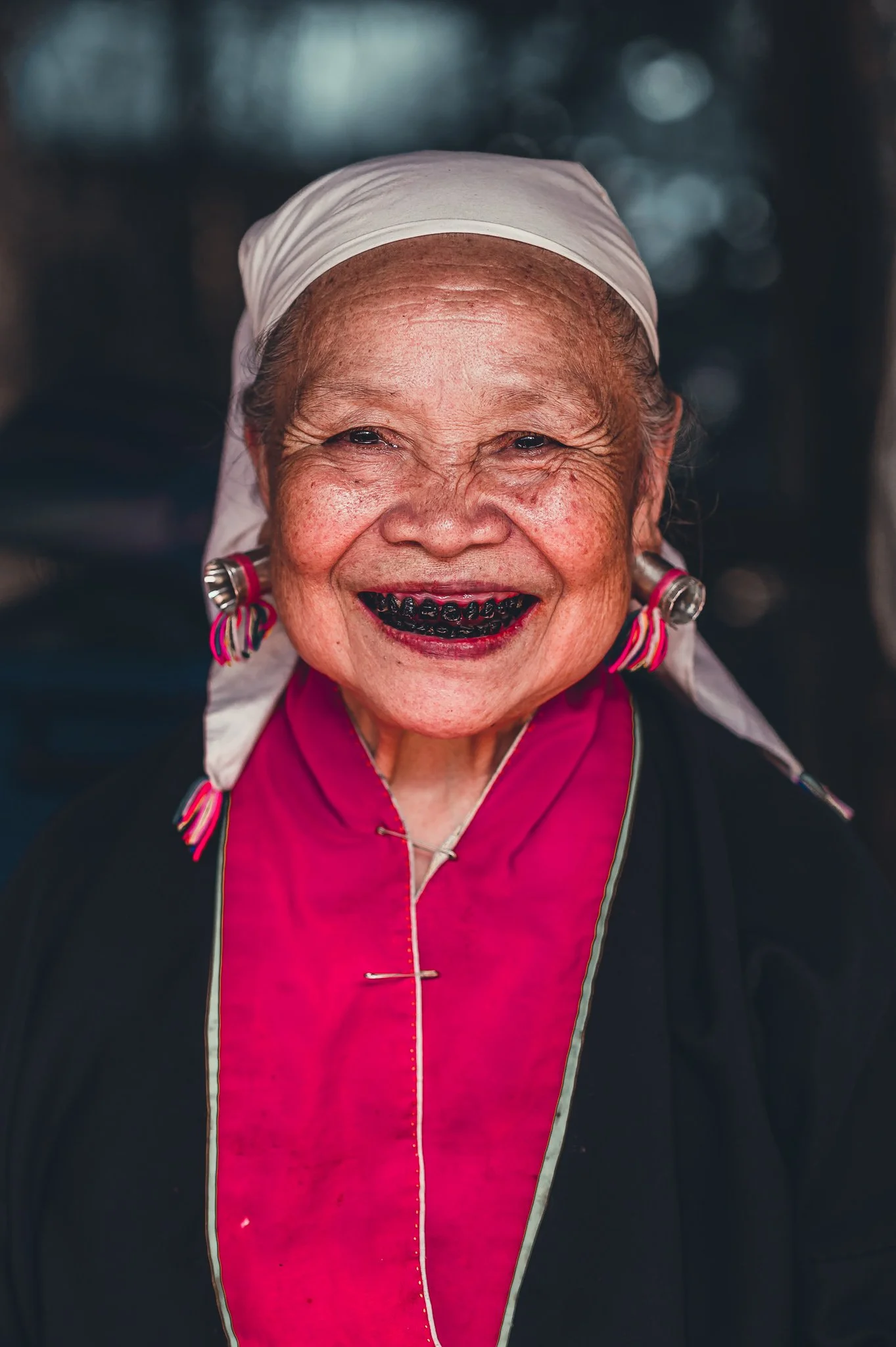 Smiling elderly woman wearing traditional clothing and headscarf, with black teeth, earrings, and a pink collar.