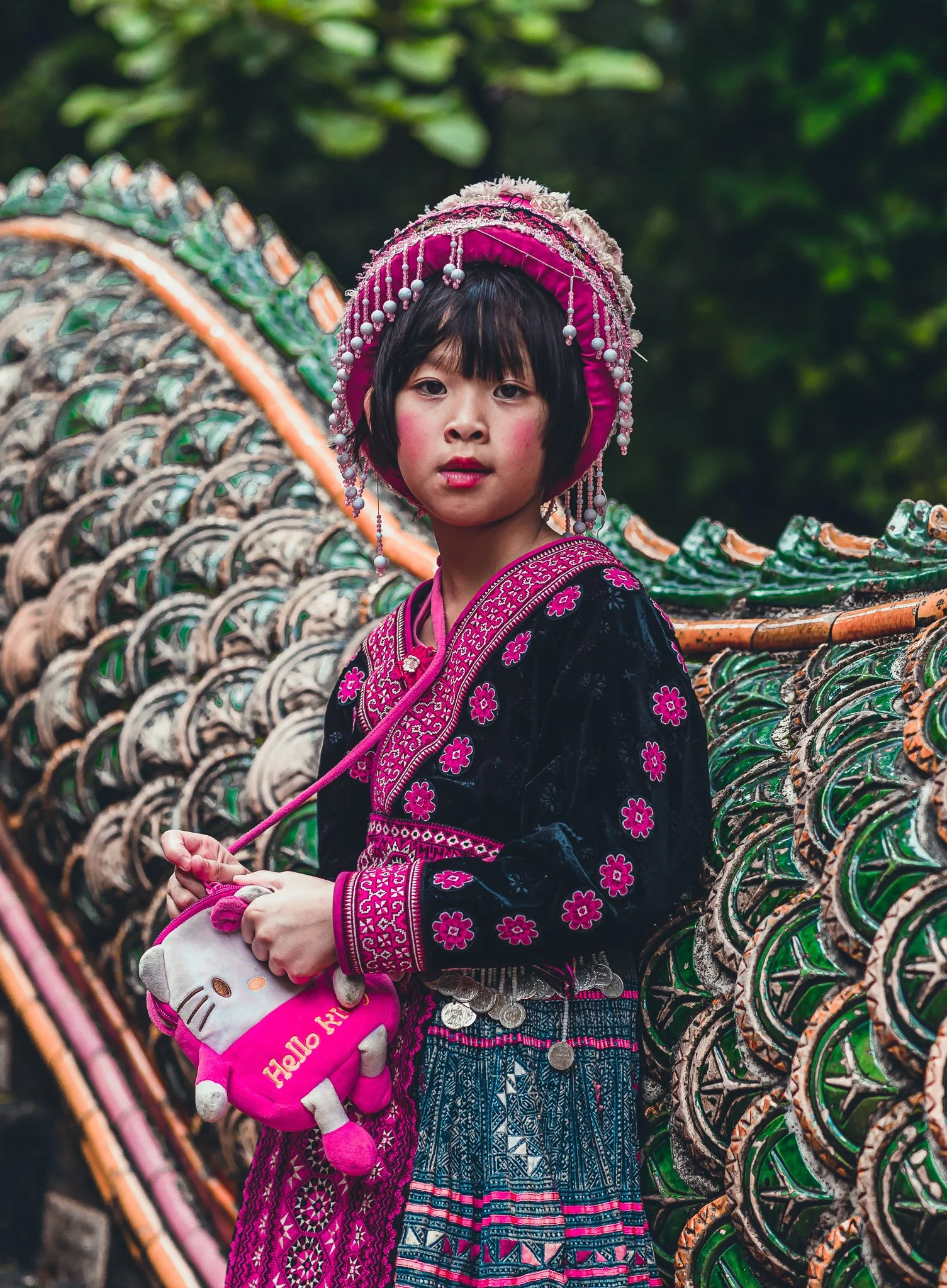 Young girl wearing traditional colorful ethnic clothing and an elaborate pink headdress, holding a Hello Kitty plush bag, standing near a decorative structure with a green and orange patterned background.