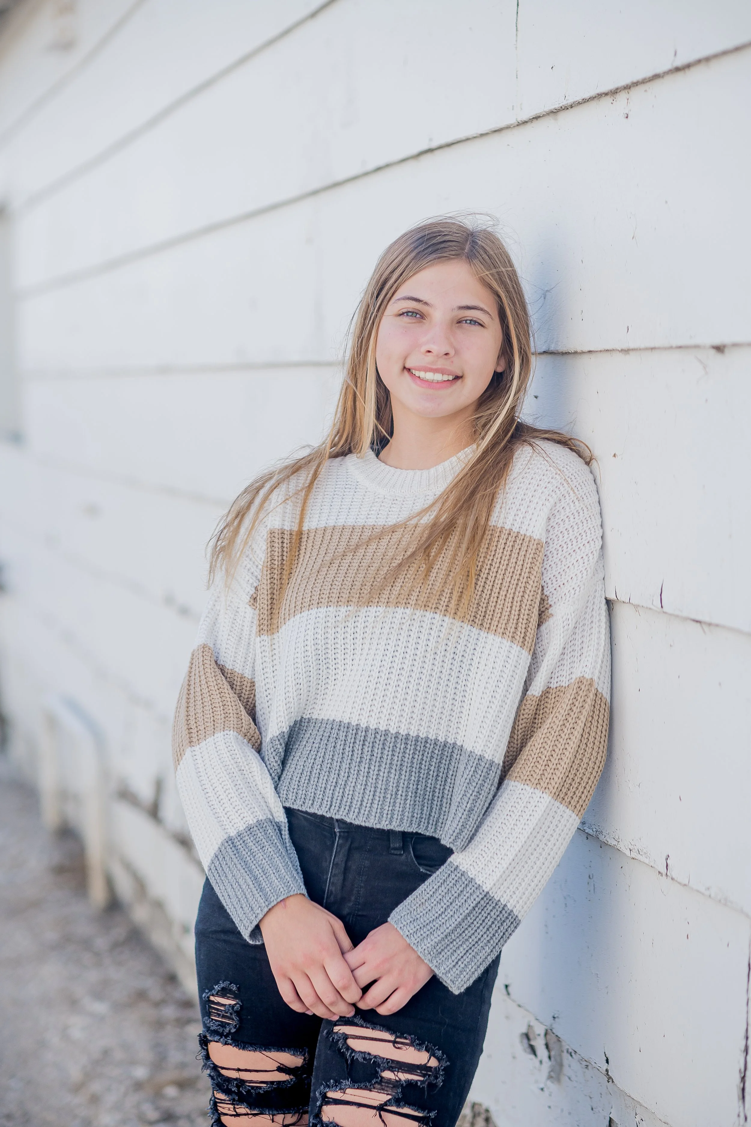 A young girl with long blonde hair smiles while leaning against a white wooden wall outdoors.