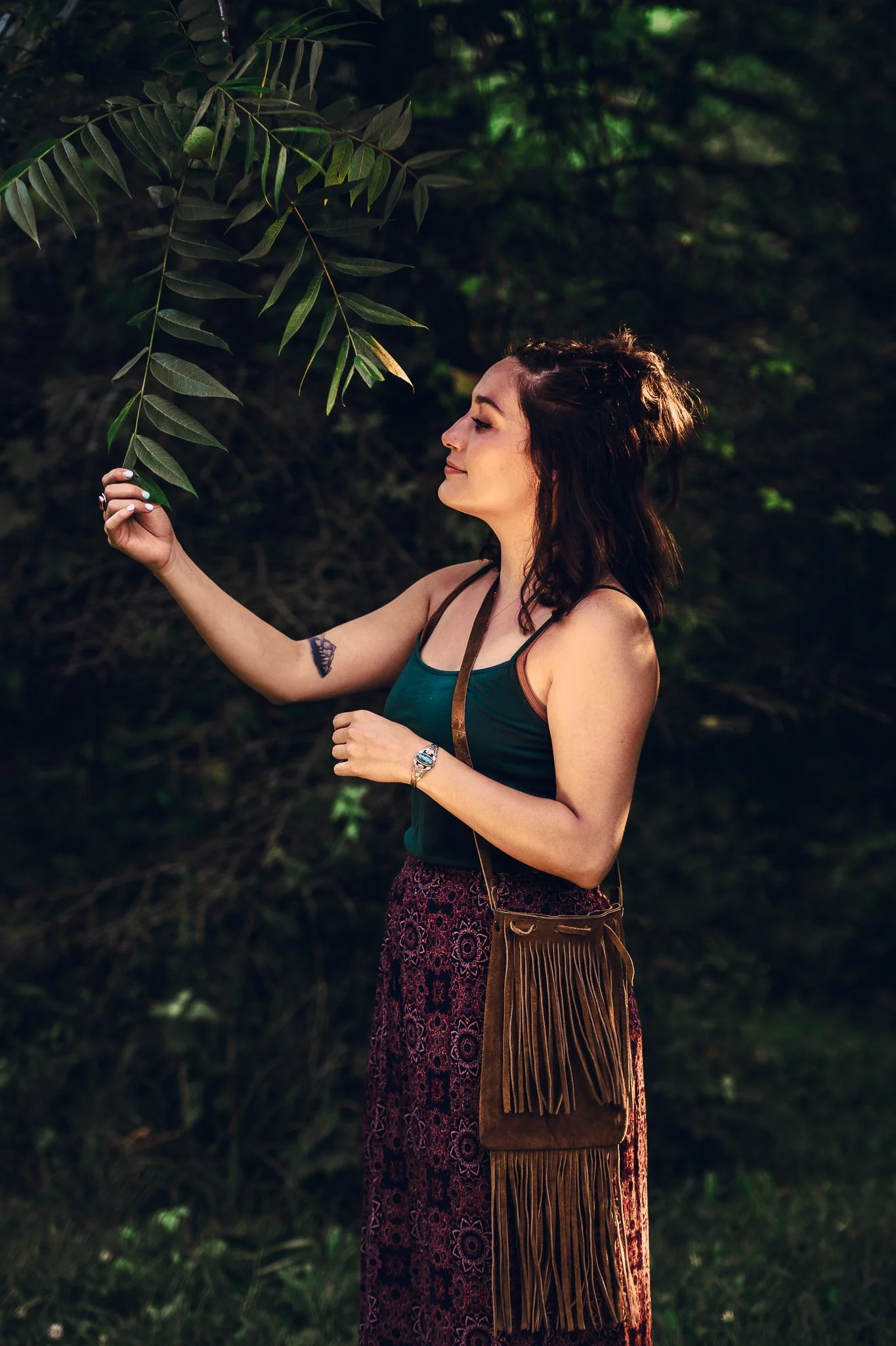 A woman in a dark green tank top and patterned skirt in a forest, holding a leaf from a tree.