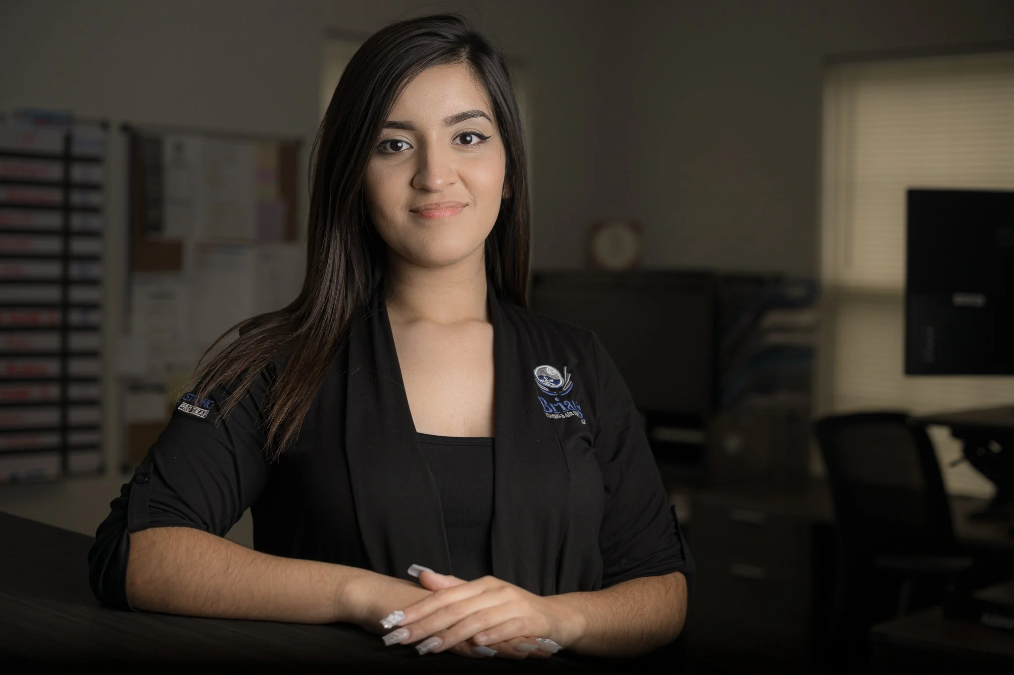 A young woman with long dark hair sitting in an office, wearing a black jacket with a logo on the chest and arm.