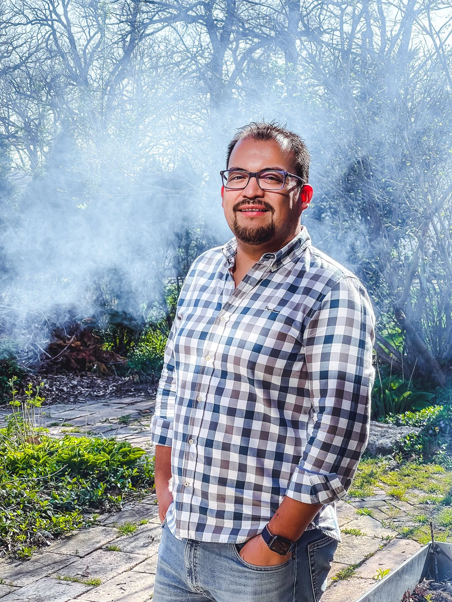 A man with glasses and a beard wearing a plaid shirt and jeans stands outdoors on a stone patio, with greenery and leafless trees in the background, and smoke or steam rising behind him.