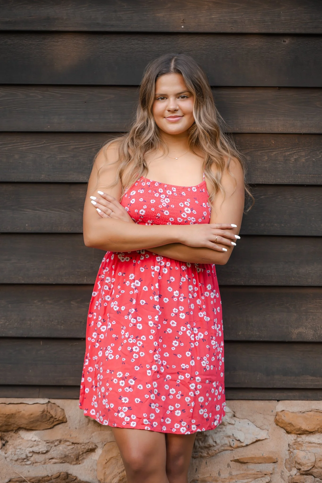 A young woman with long wavy hair, wearing a red floral dress, standing with arms crossed in front of a dark wooden wall.