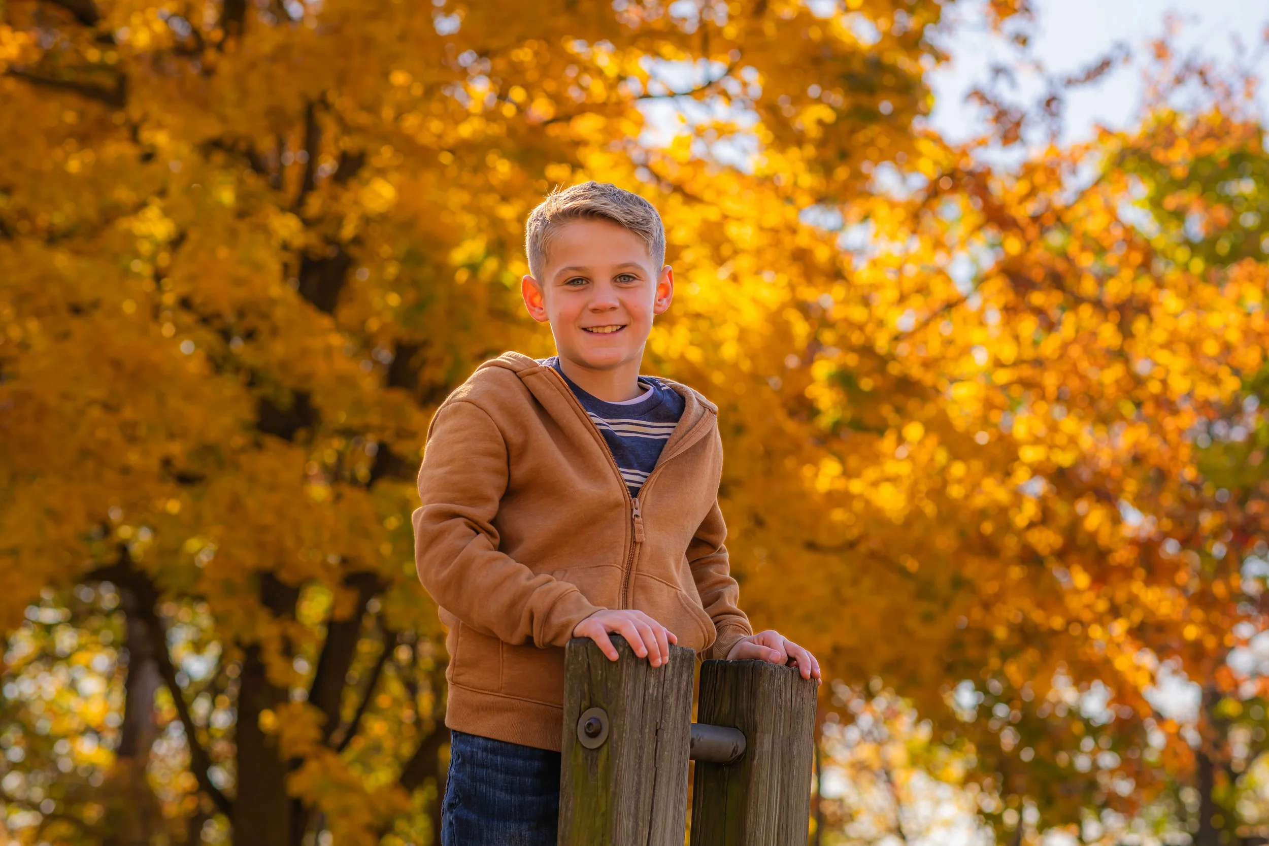 A young boy with blond hair, smiling, wearing a brown hoodie and striped shirt, standing outdoors among trees with vibrant orange and yellow autumn leaves.