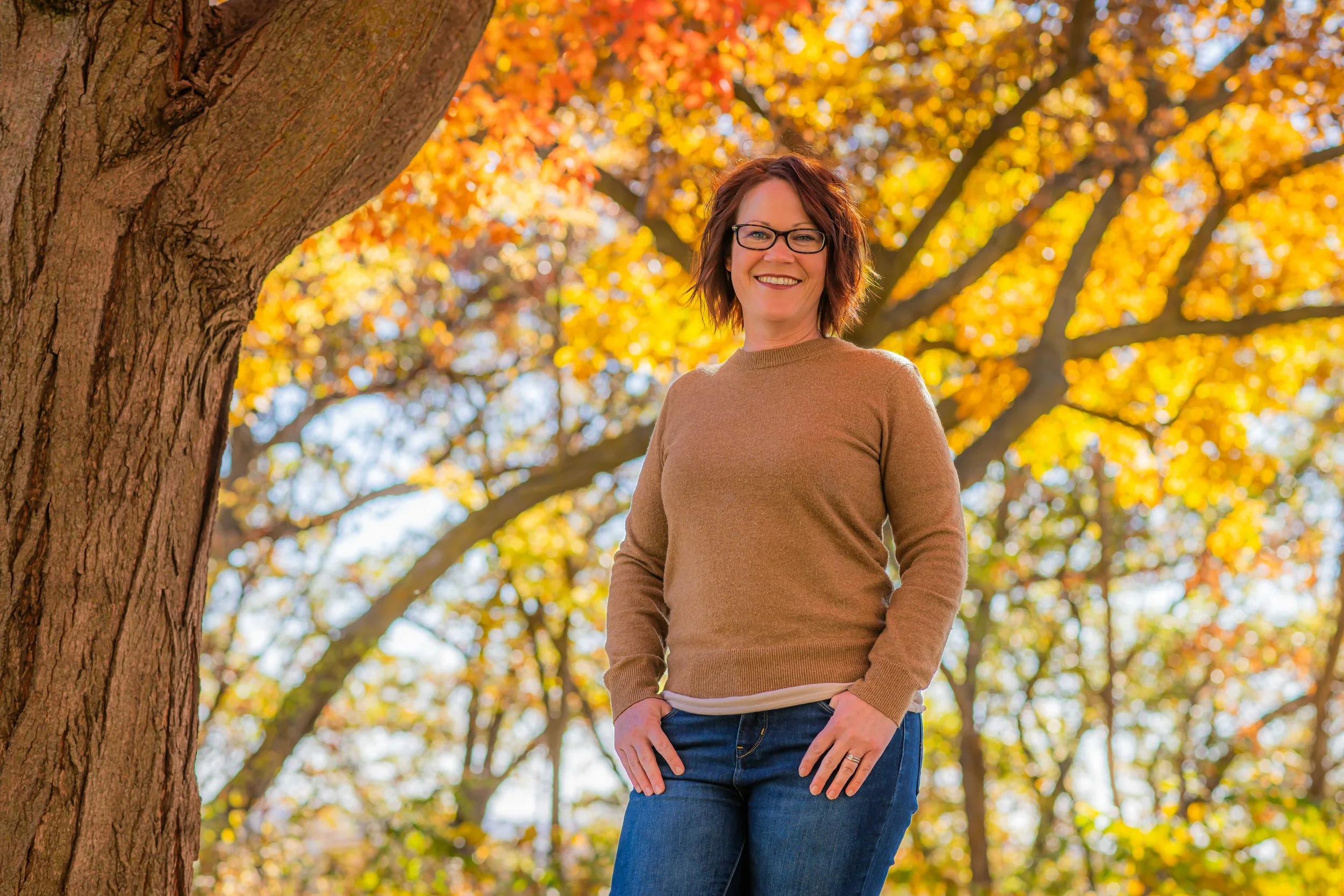 A woman with red hair, glasses, and a warm smile standing outdoors near a large tree in a park with autumn-colored leaves.