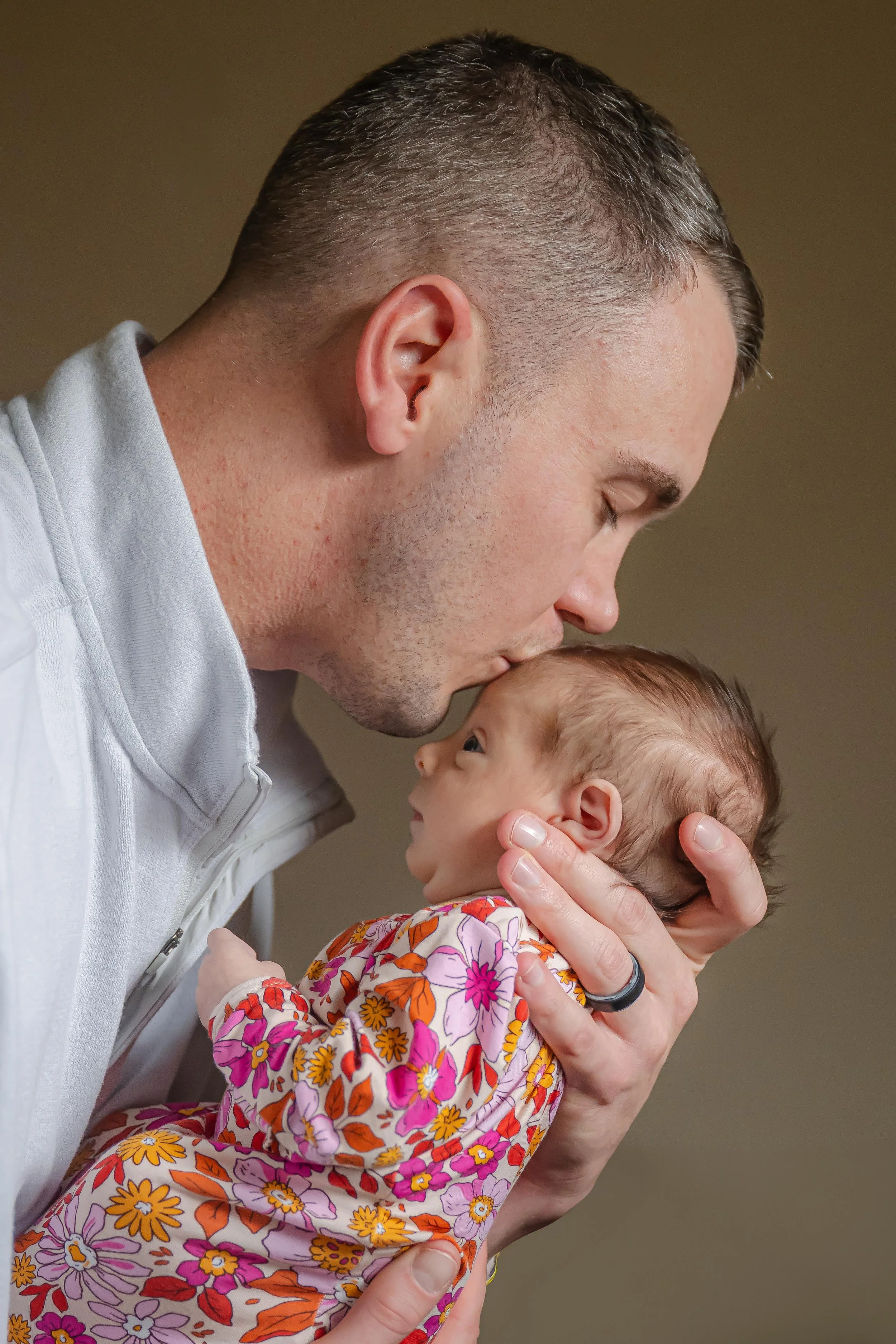A man with short hair and a gray shirt gently kisses a newborn baby on the forehead, holding the baby close with a floral outfit, against a plain background.