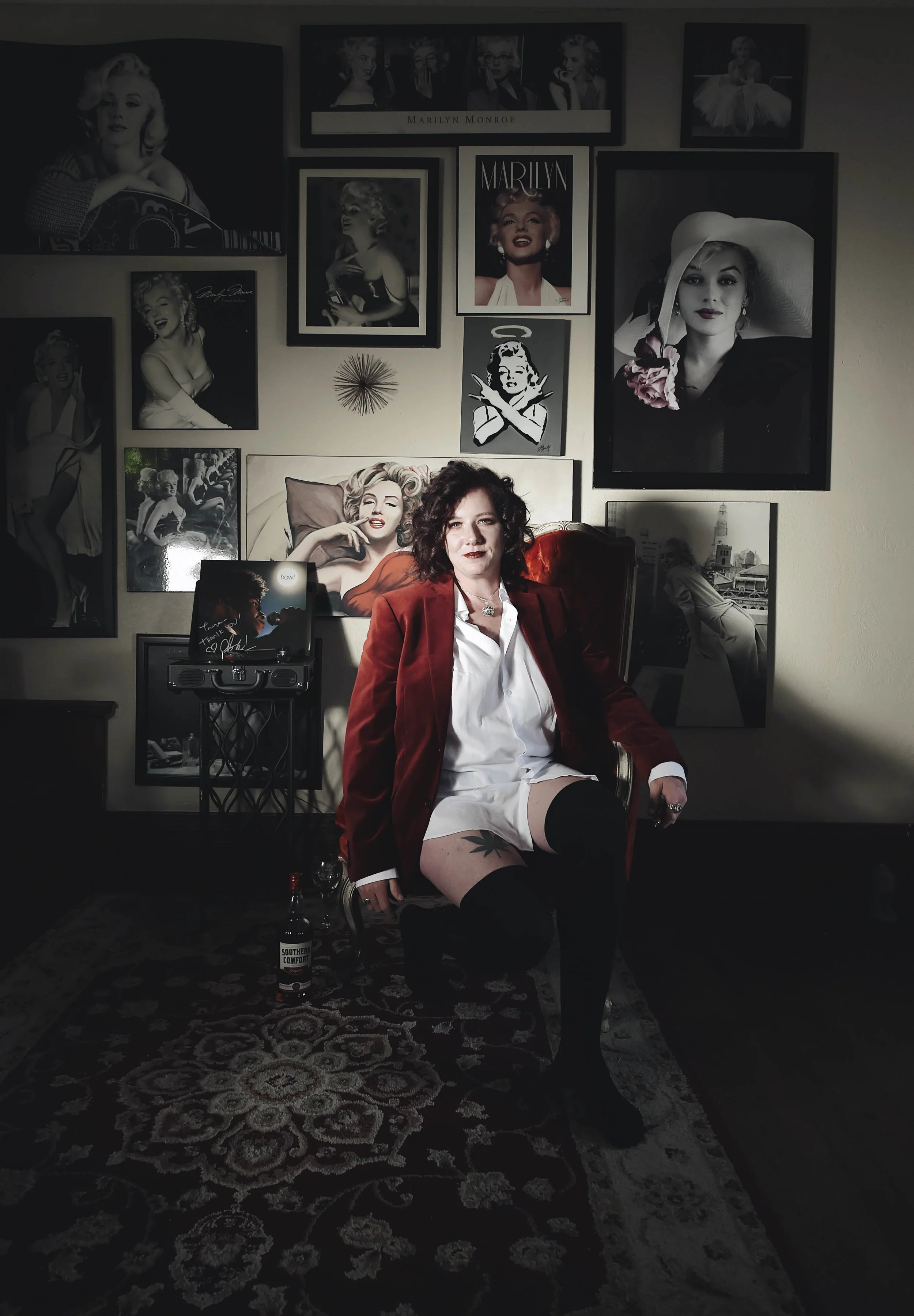 Woman with curly hair in a red blazer and white shirt seated in a vintage chair in front of a wall decorated with Marilyn Monroe posters and photographs. She wears thigh-high stockings, and a tattoo is visible on her thigh. There is a bottle of whisk