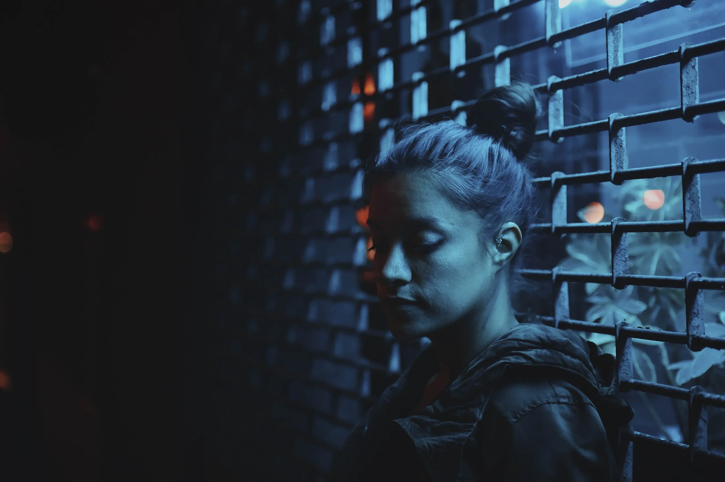 A woman with her hair in a bun, wearing a jacket, standing against a brick wall with blue lighting and some blurred orange lights in the background.