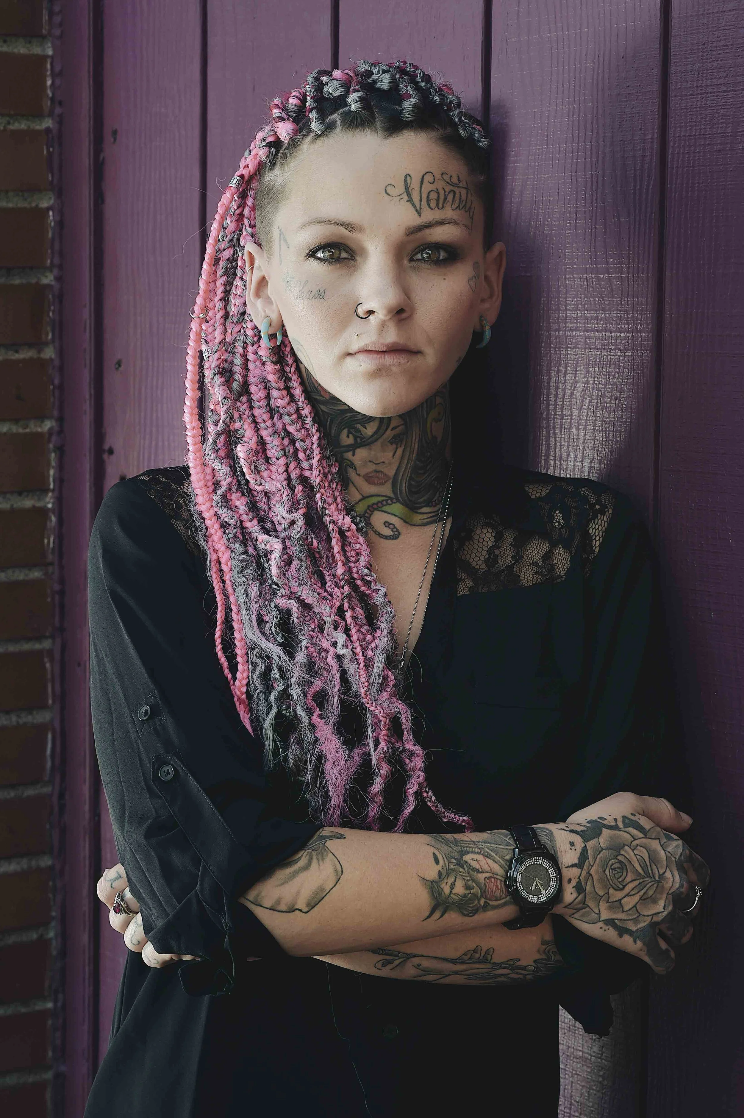 Portrait of a woman with pink and black dreadlocks, tattoos on her face, neck, and arms, wearing a black lace top, standing in front of purple wooden paneling.