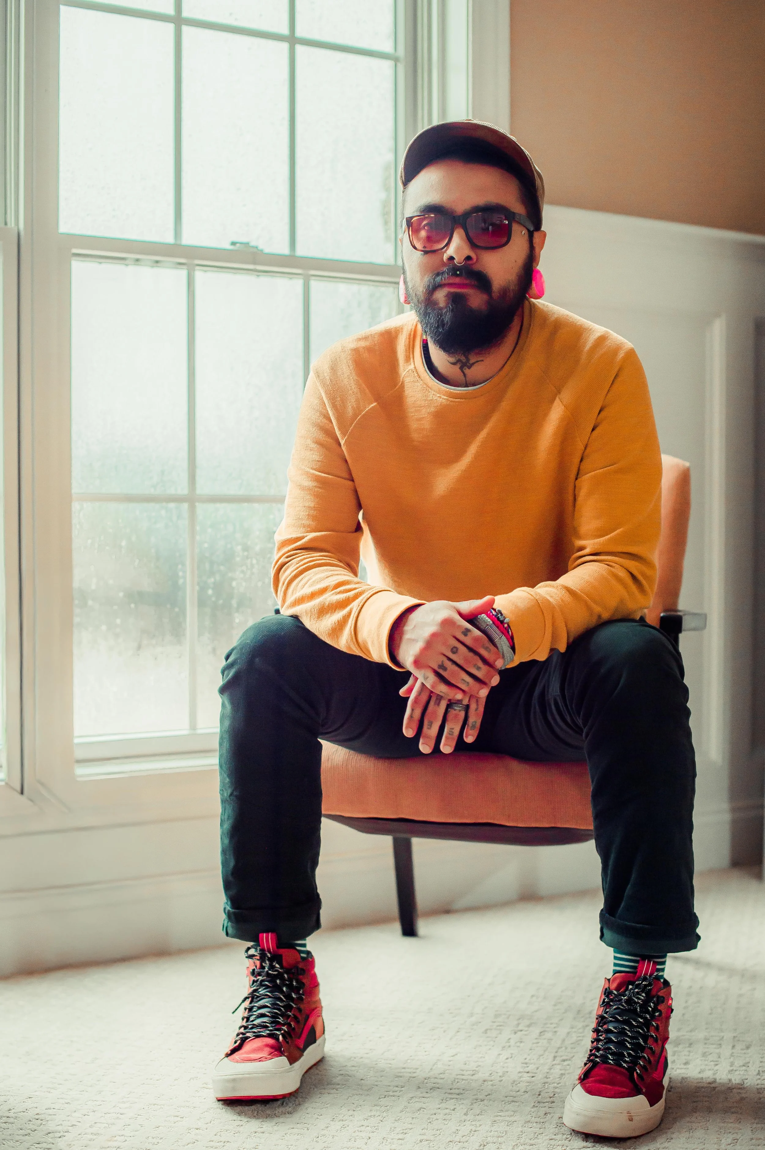 A man with a beard, tattoos, and wearing sunglasses, a yellow sweatshirt, black pants, and red high-top sneakers, sitting in a room with large windows and beige walls.