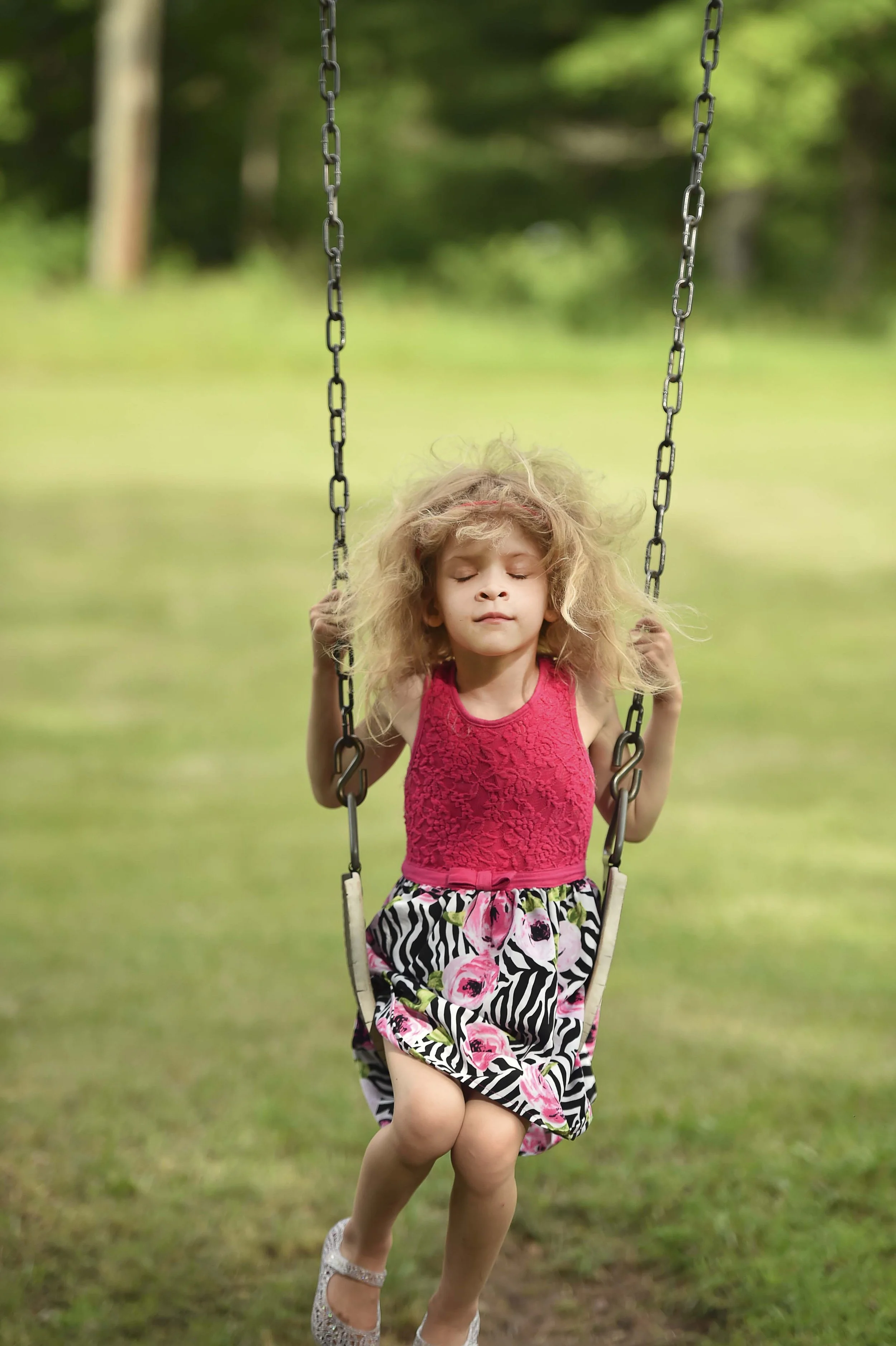 Young girl with curly blonde hair and closed eyes sitting on a swing outdoors in a grassy park.