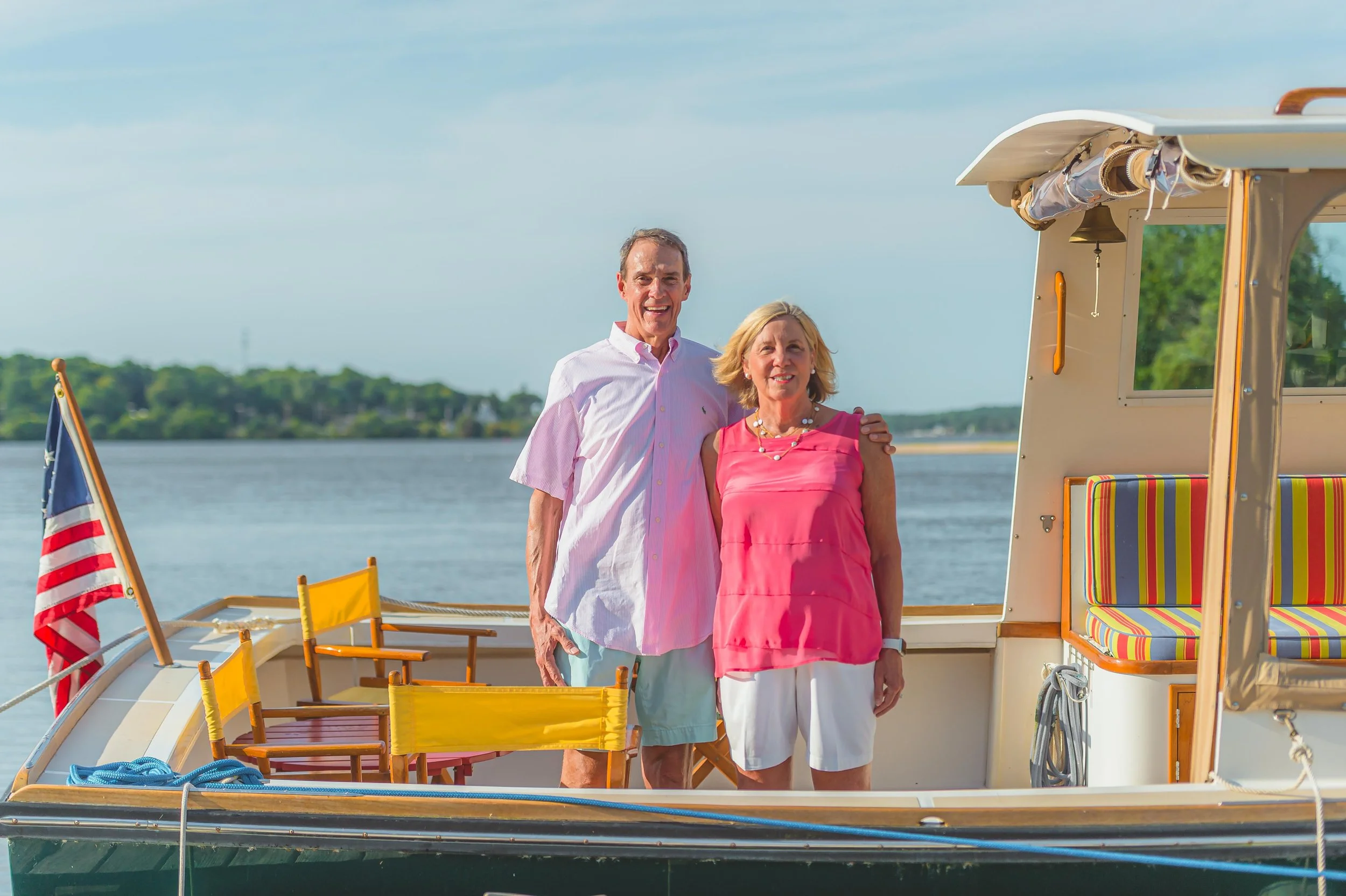 A smiling couple standing on a boat near a body of water with a green shoreline in the background.