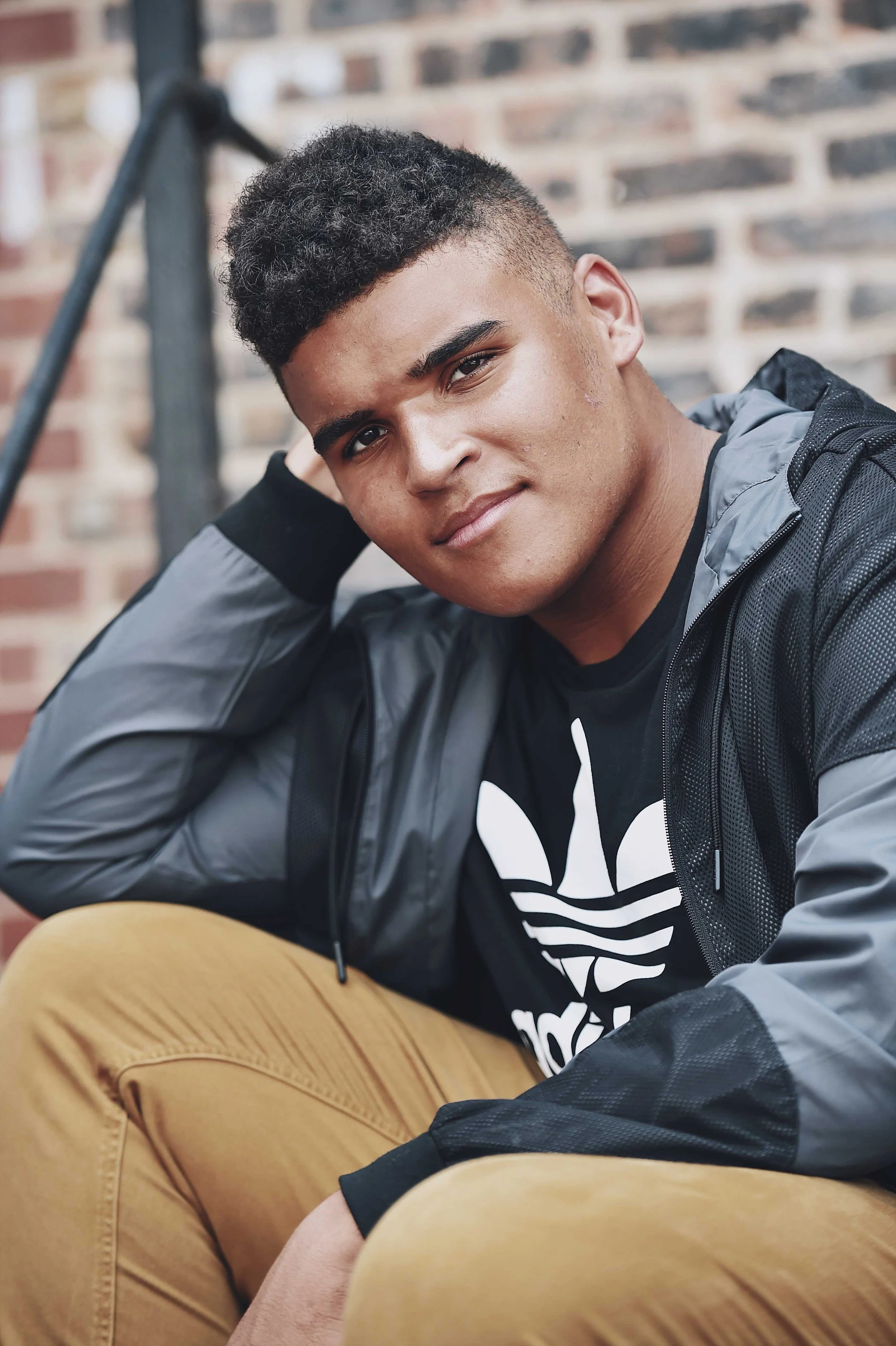 A young man with curly hair and a dark skin tone, sitting outdoors against a brick wall, casually posing with one arm resting on his knee and the other hand behind his head. He wears a black T-shirt with a white Adidas logo and a black and gray jacke
