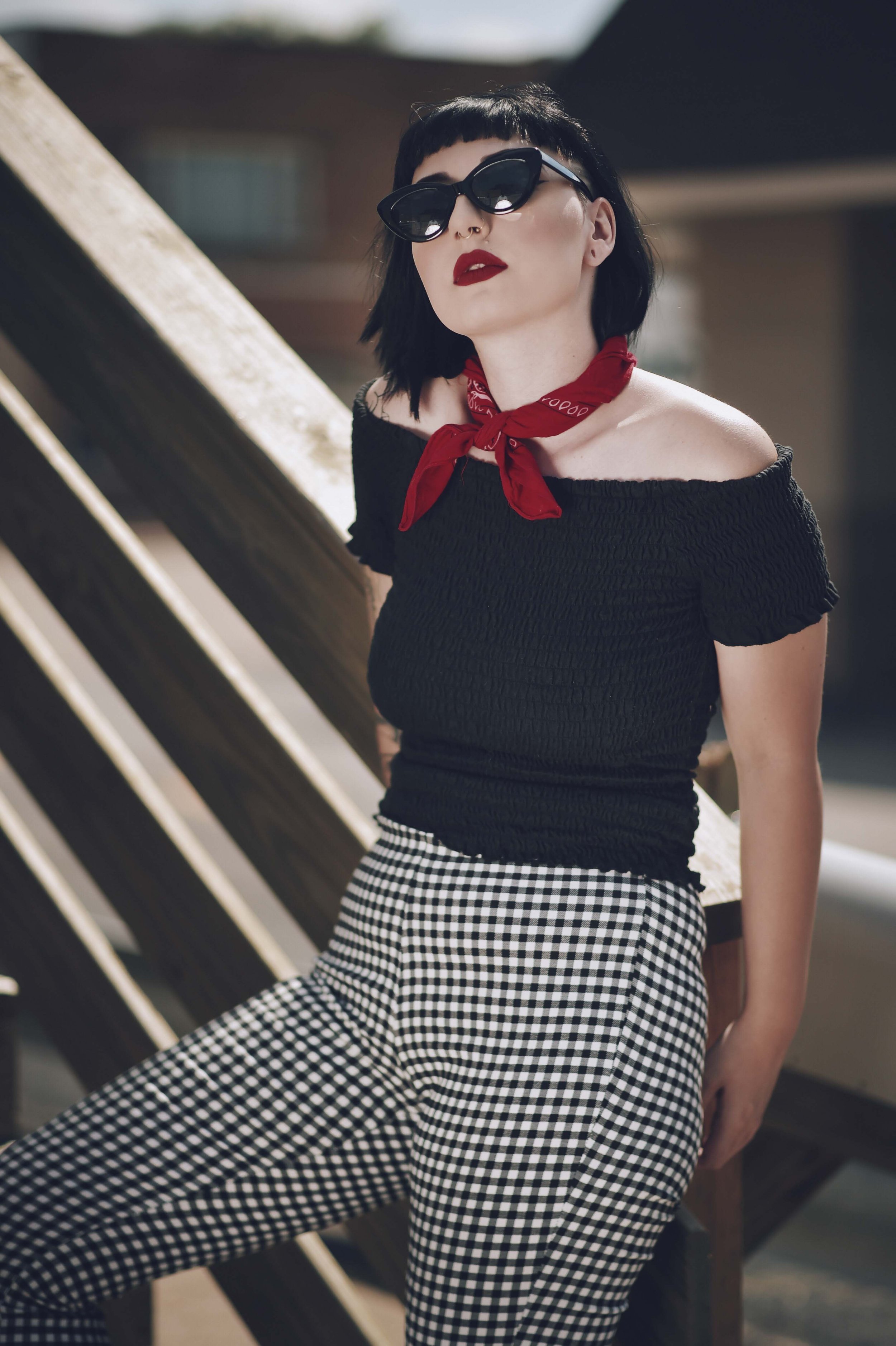 Woman with dark hair, sunglasses, and red bandana around her neck, wearing a black off-the-shoulder top and black-and-white checkered pants, leaning against a wooden railing outdoors.
