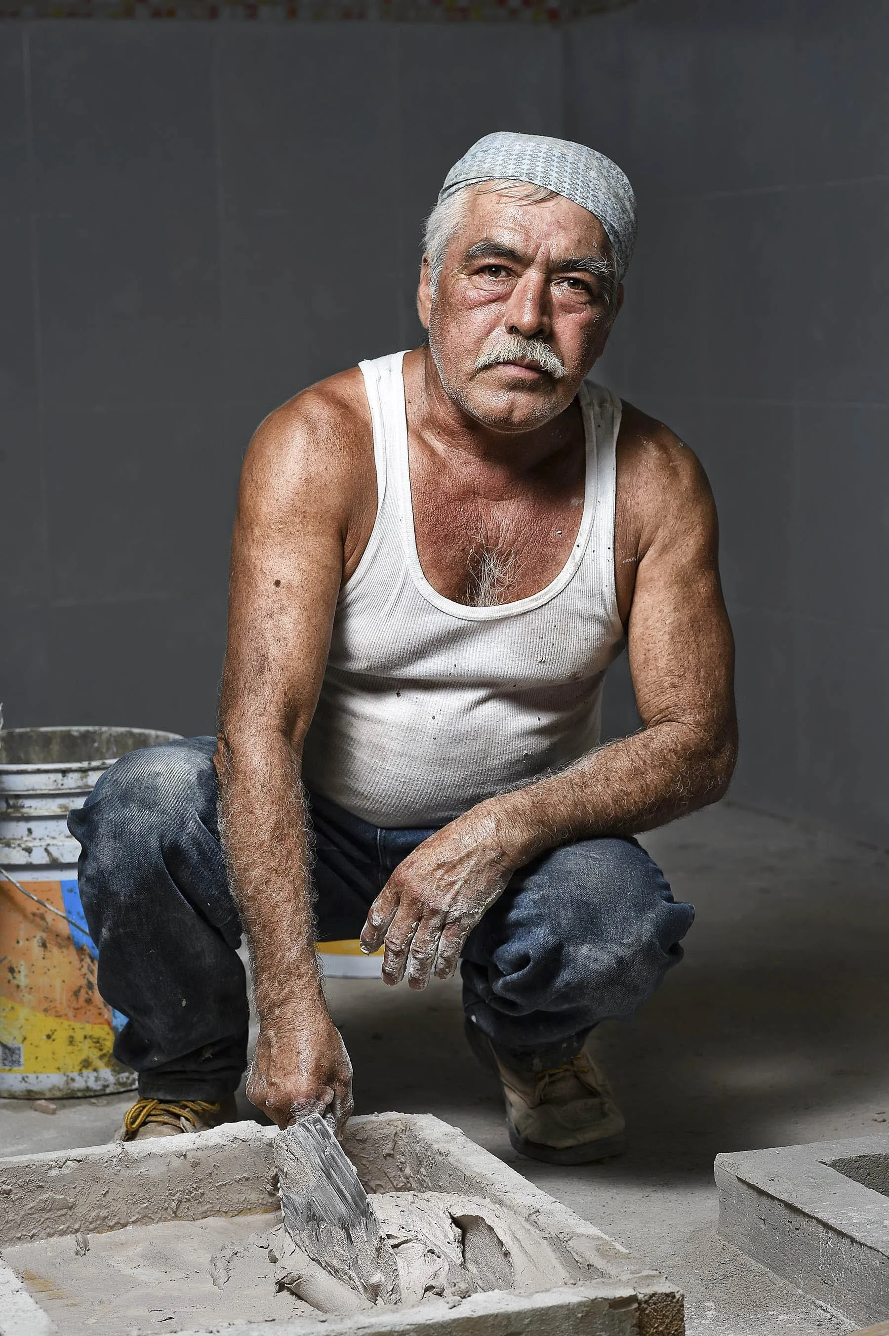 An elderly man with gray hair, mustache, and beard, wearing a white tank top, kneeling on the ground, working with cement or plaster, with a trowel in his hand.