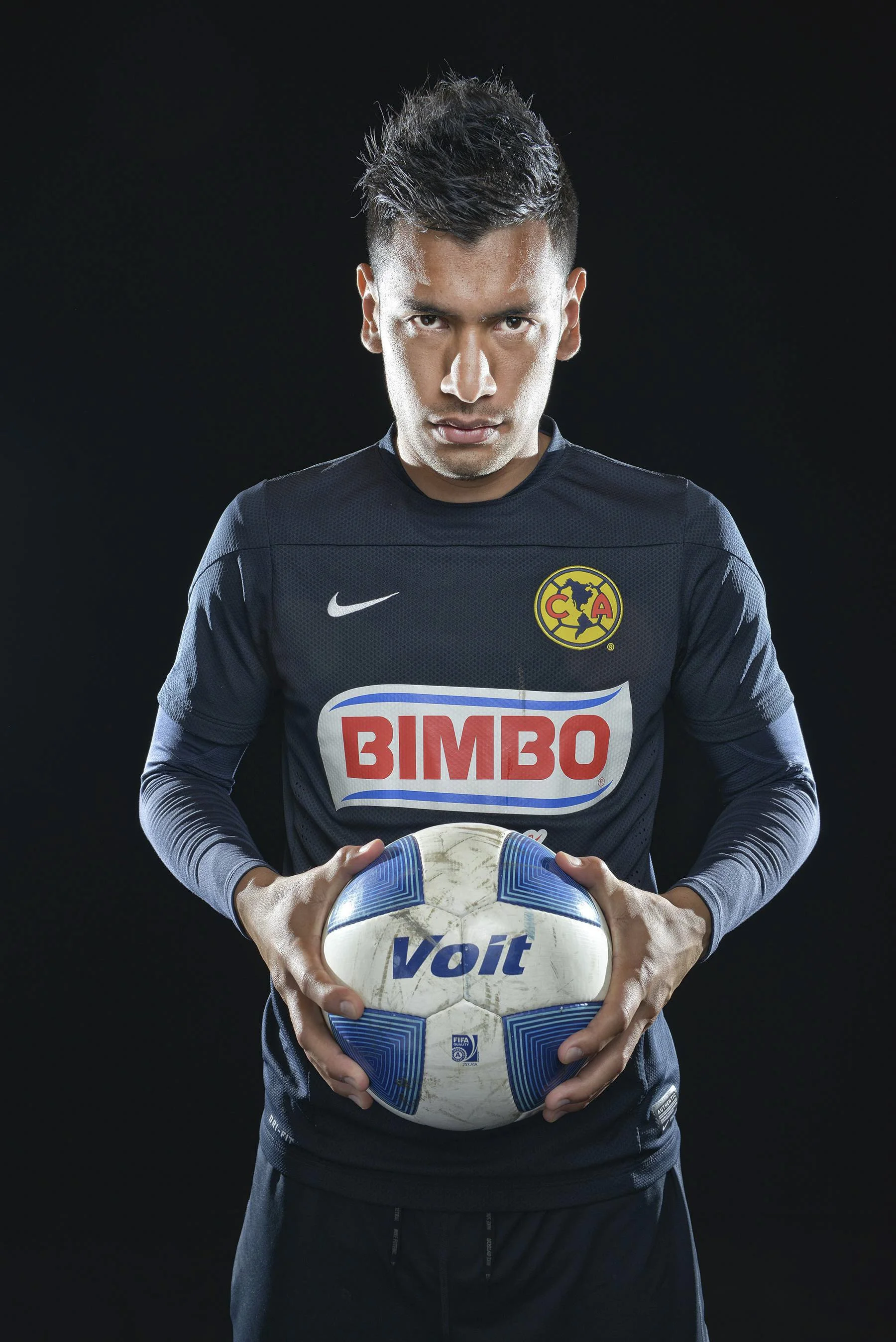 A male soccer player in a dark blue jersey holding a soccer ball in front of him against a black background.