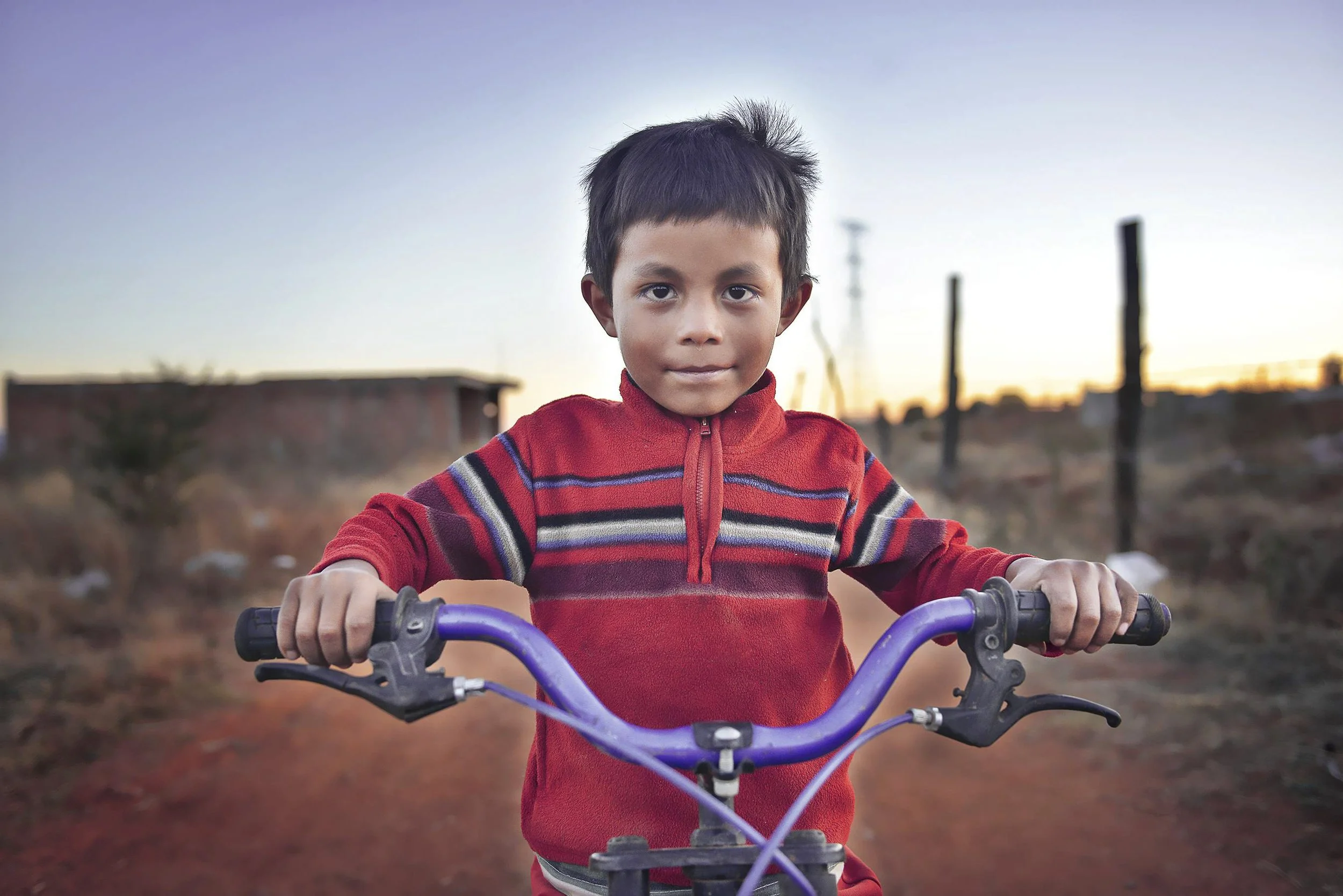 Young boy riding a bicycle outdoors during sunset, wearing a red sweater with stripes.