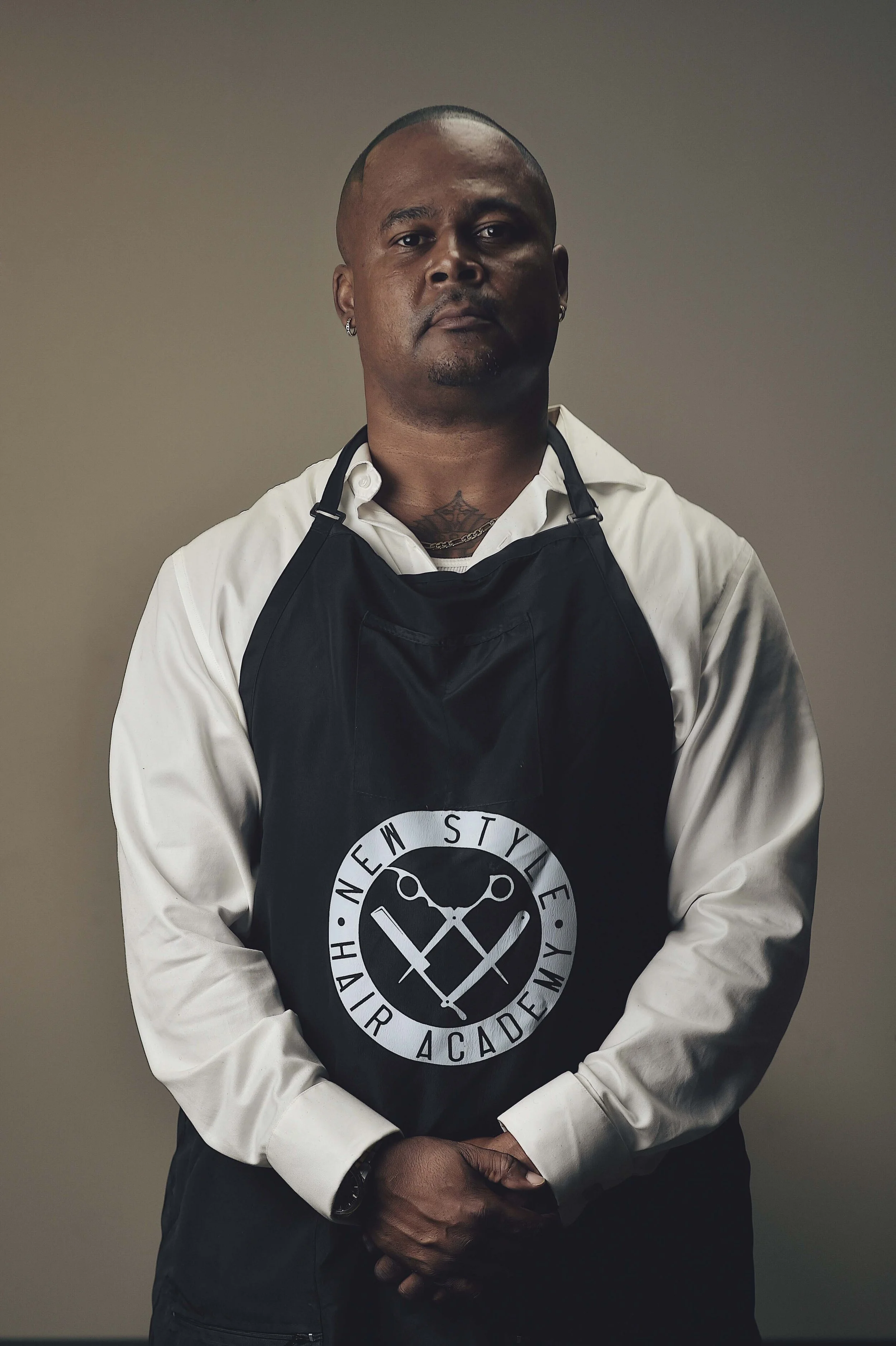 A man wearing a white shirt and a black apron with the logo 'New Style Hair Barber Academy' stands against a plain background.