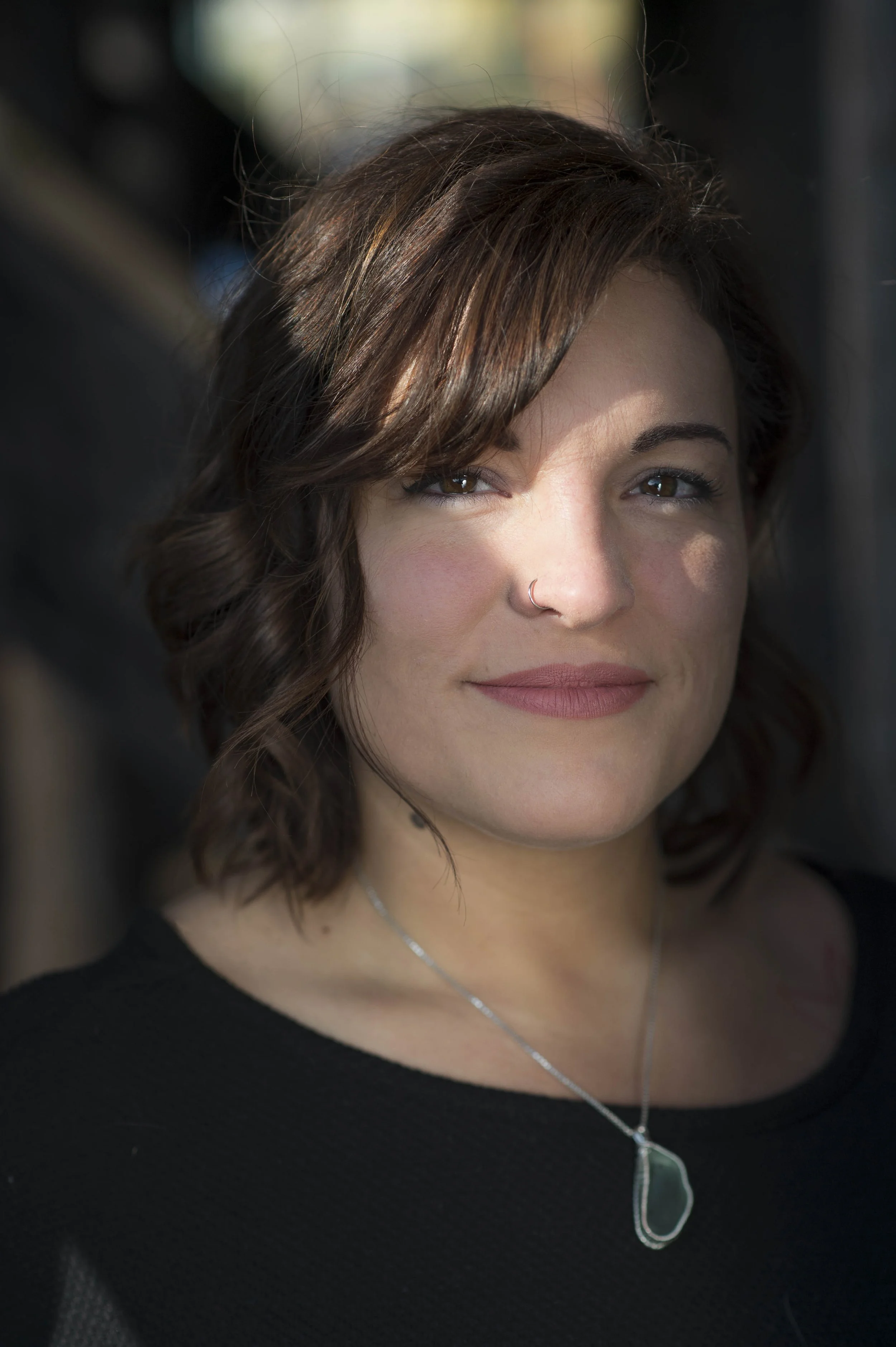 A woman with shoulder-length wavy brown hair, light skin, and brown eyes looks at the camera with a subtle smile. She wears a nose ring, a silver necklace with a green stone pendant, and a black top.