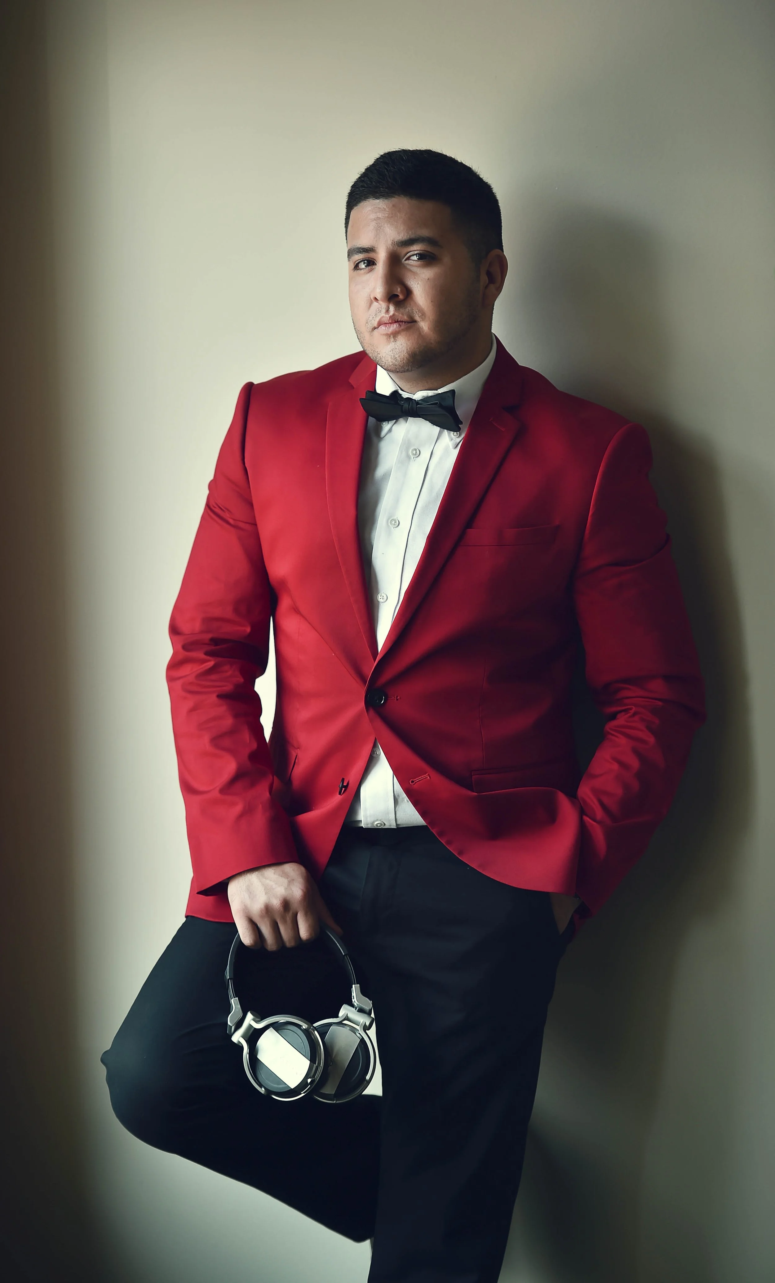 A man in a red tuxedo jacket, white dress shirt, and black bow tie, holding a pair of headphones and standing against a plain wall.